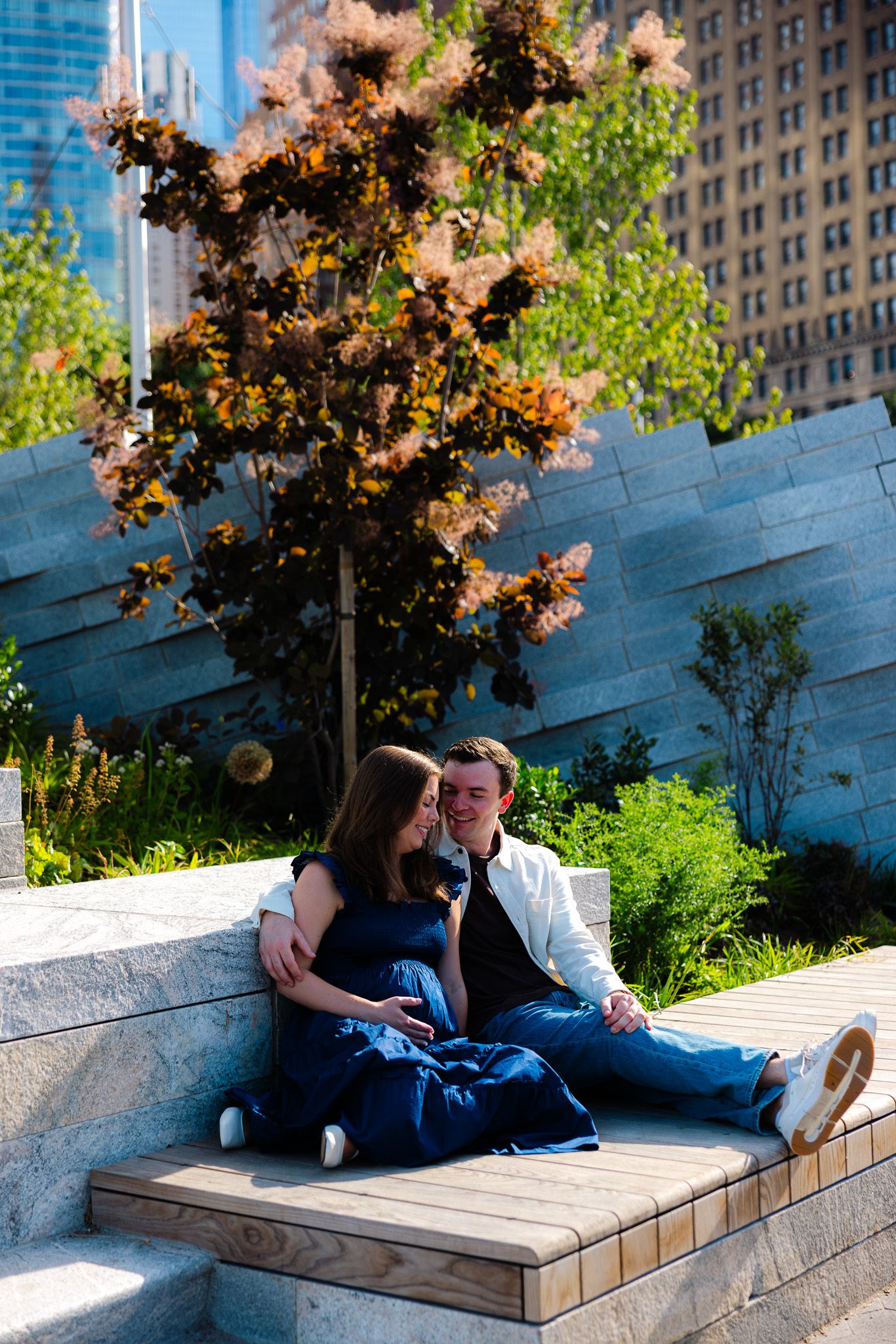 Couple sitting together on stone steps in an urban garden, surrounded by lush greenery and colorful trees.