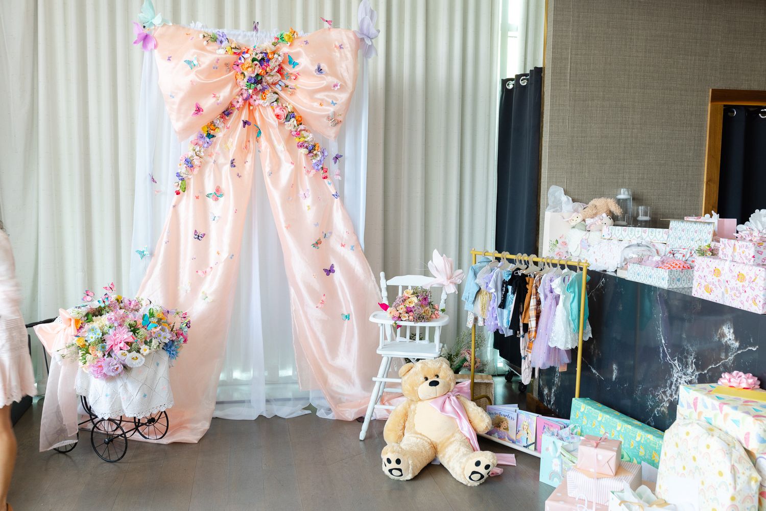 Pink draped baby shower backdrop with floral bow, teddy bear, vintage pram, and colorful baby clothes display.