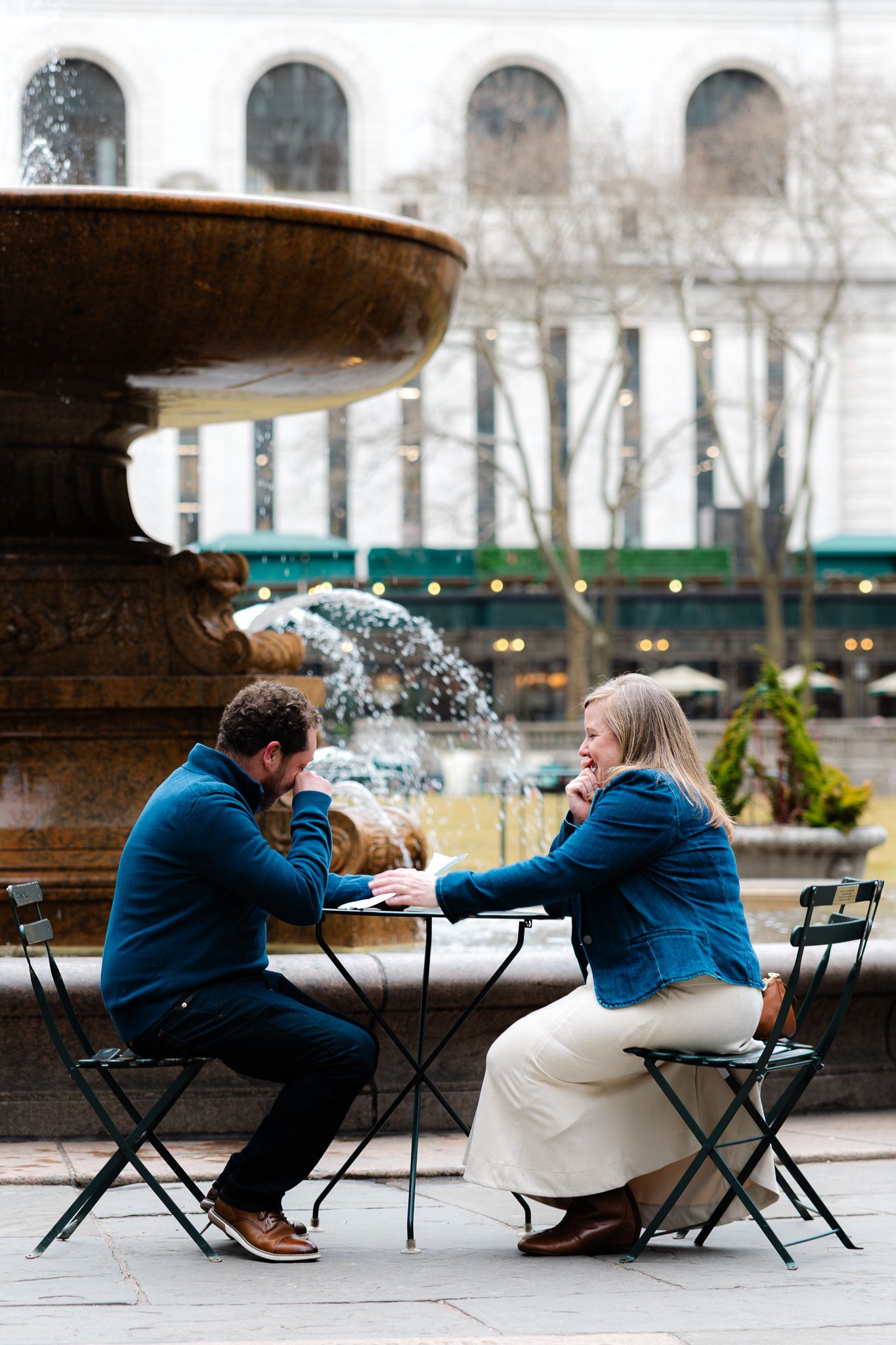 Couple in matching blue jackets sit at outdoor cafe table by ornate stone fountain, engaged in conversation.
