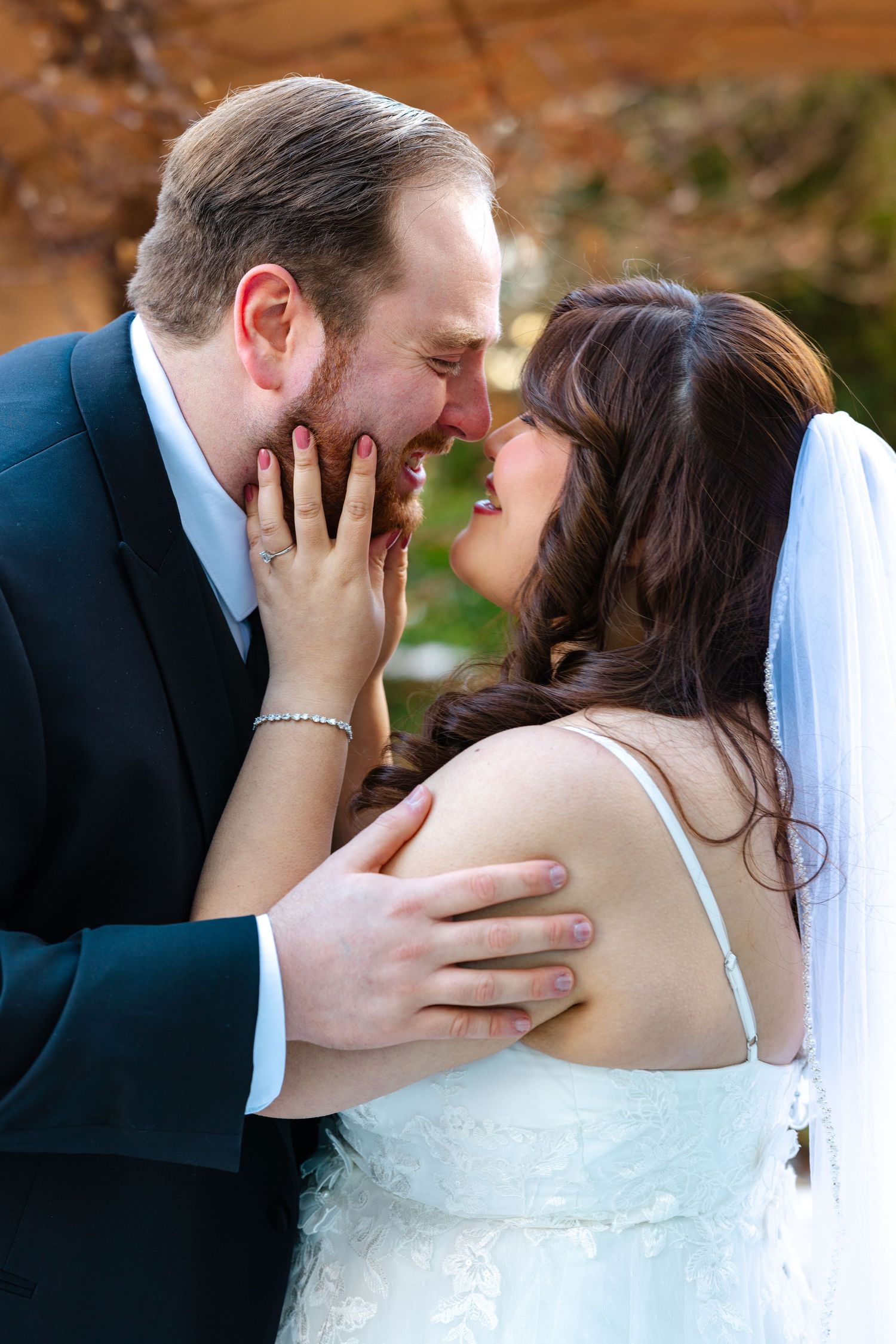 Groom whispers to laughing bride in lace gown during romantic outdoor wedding portrait.