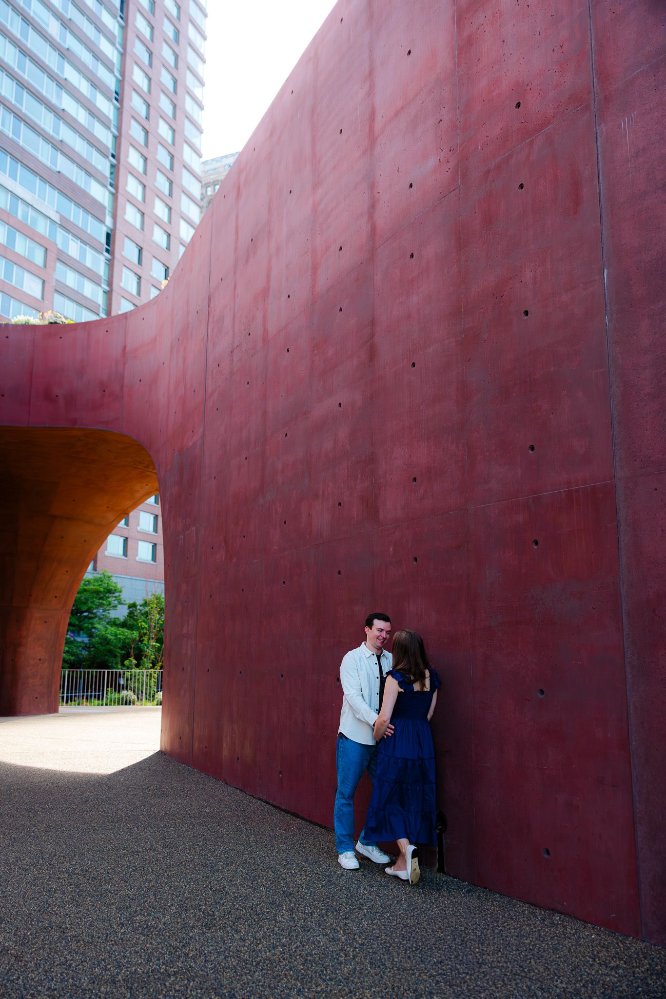 Couple sharing a romantic moment leaning against a curved red concrete wall in an urban setting.