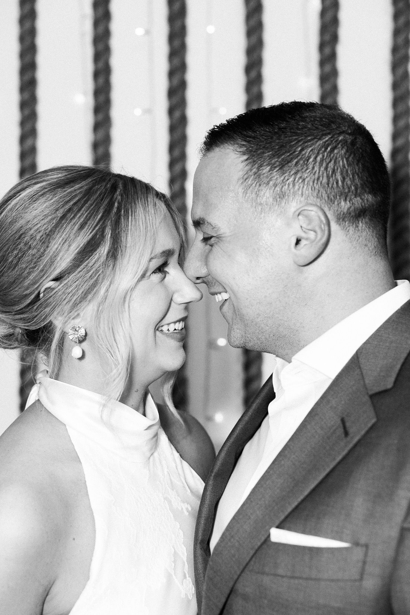 Black and white photo of a smiling couple in formal attire gazing at each other at a wedding celebration.