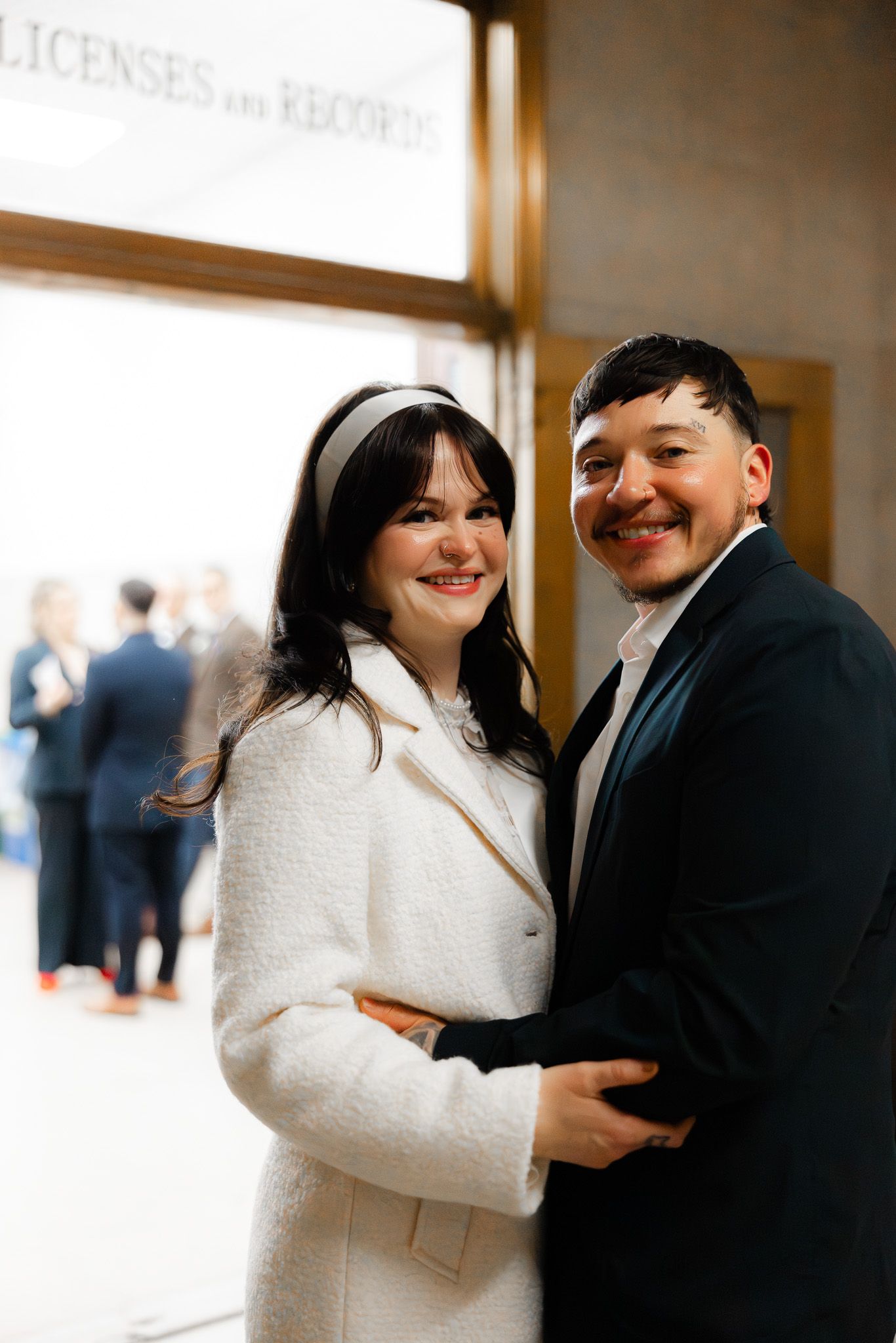 Smiling couple embracing in front of Licenses and Records office, bride in white coat with headband, groom in dark suit.