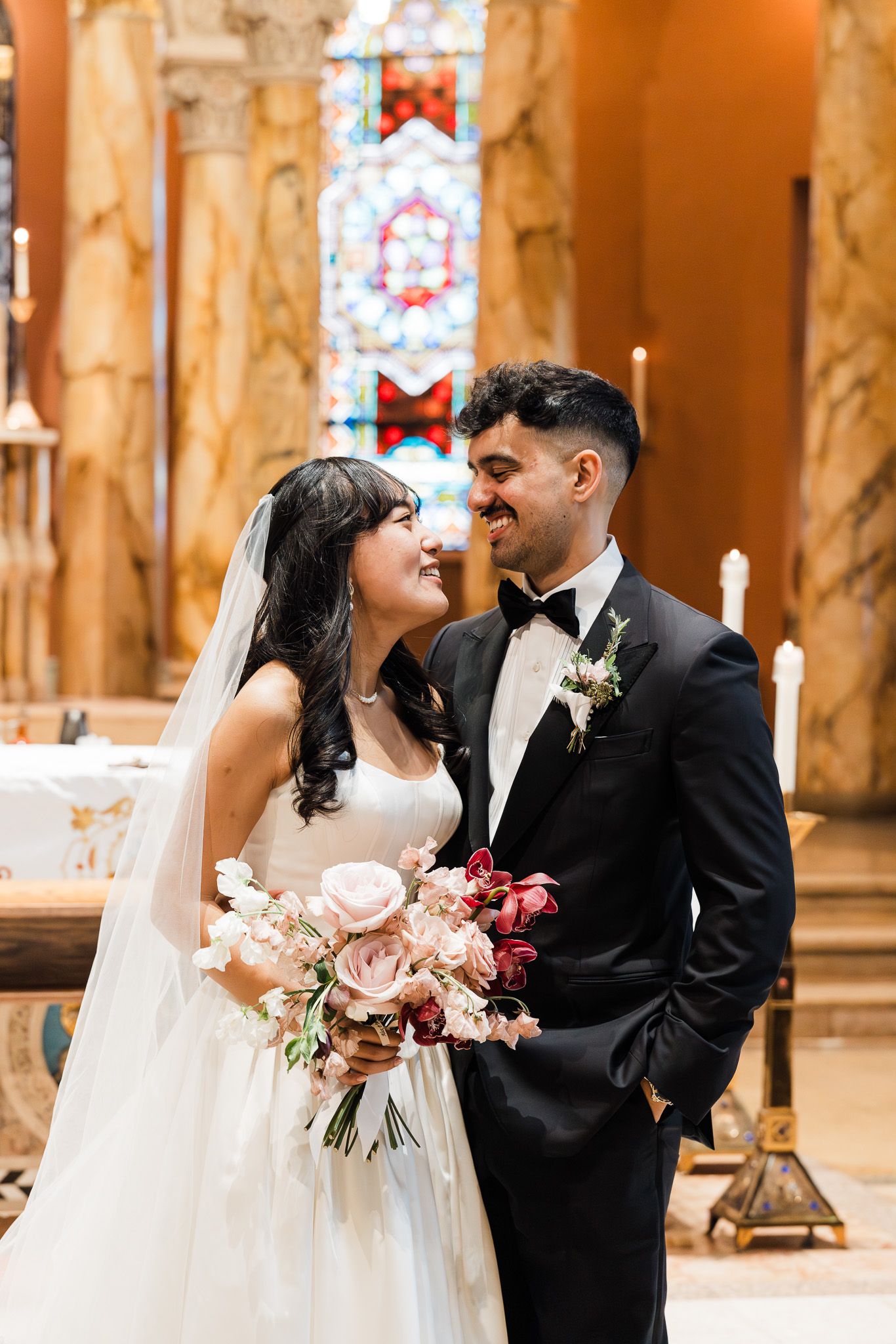 Bride and groom smiling at each other inside an ornate church with stained glass windows.