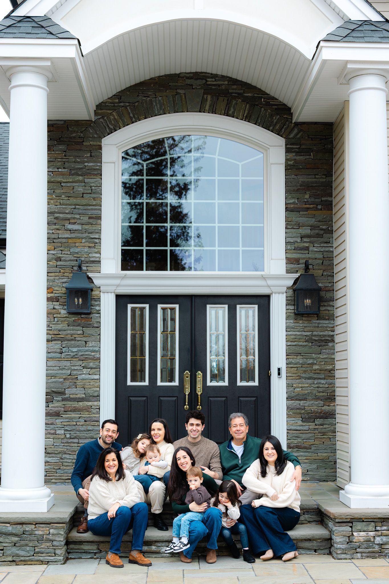 Large family group portrait posed on front steps of elegant stone home with arched window and white columns.