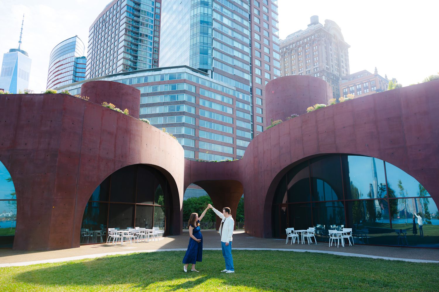 Couple dancing outdoors near modern red arch sculpture with NYC skyline and skyscrapers in background.