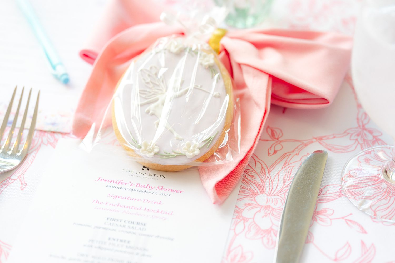 Elegant baby shower cookie with white icing and pink bow on a floral menu card, surrounded by silverware.