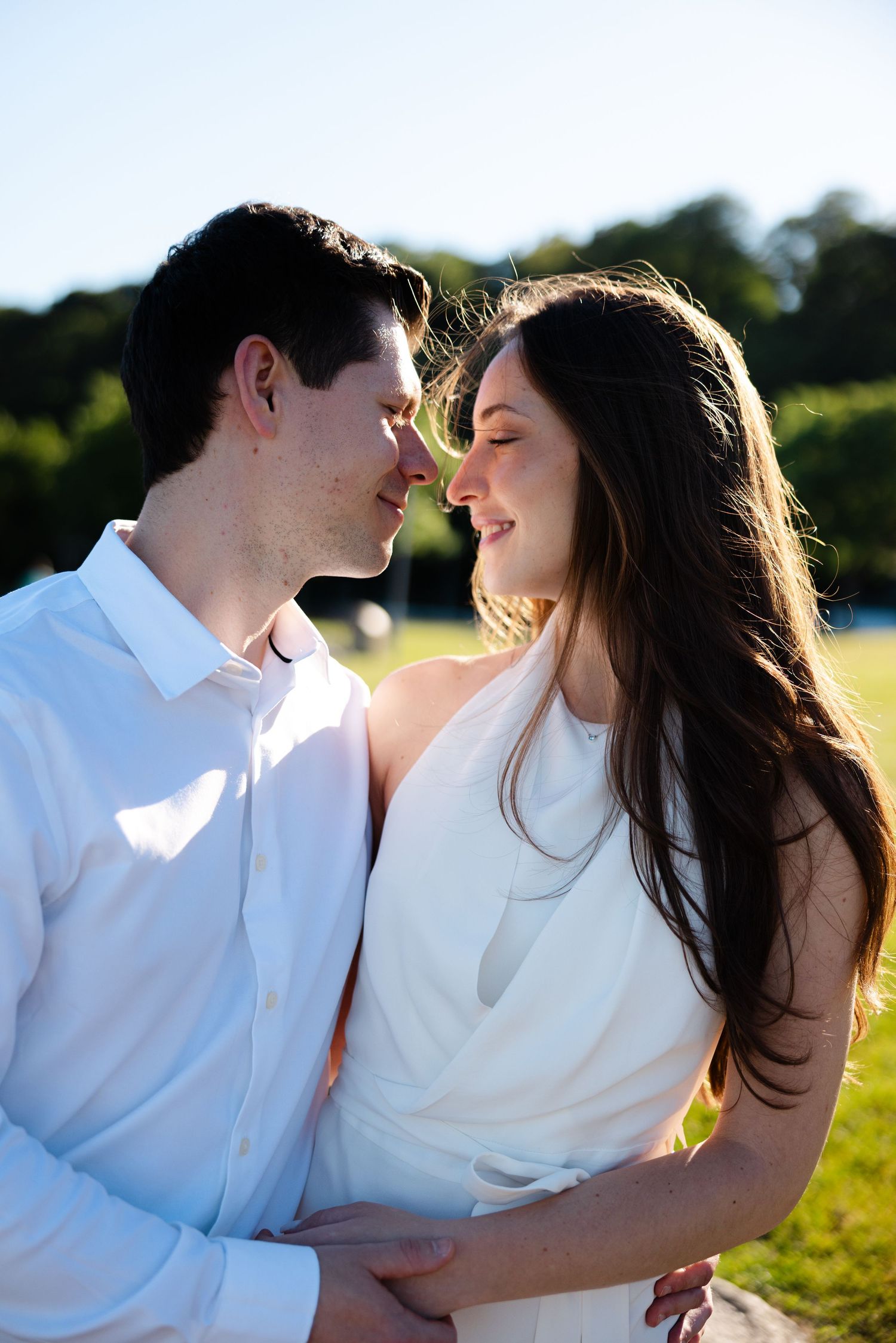 Couple in white attire sharing a romantic moment outdoors, smiling at each other in warm sunlight.
