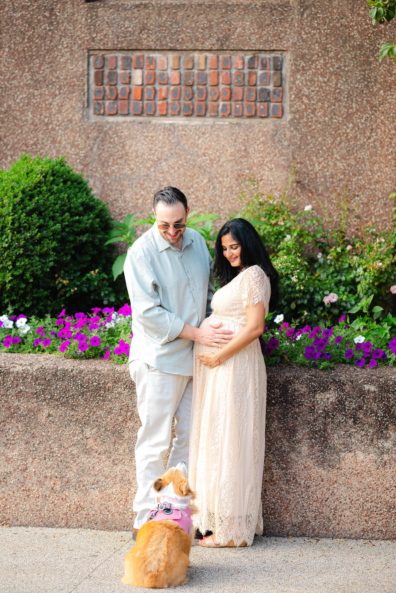 Couple in garden setting, pregnant woman in white lace dress, man in light blue shirt, golden dog with pink bow.