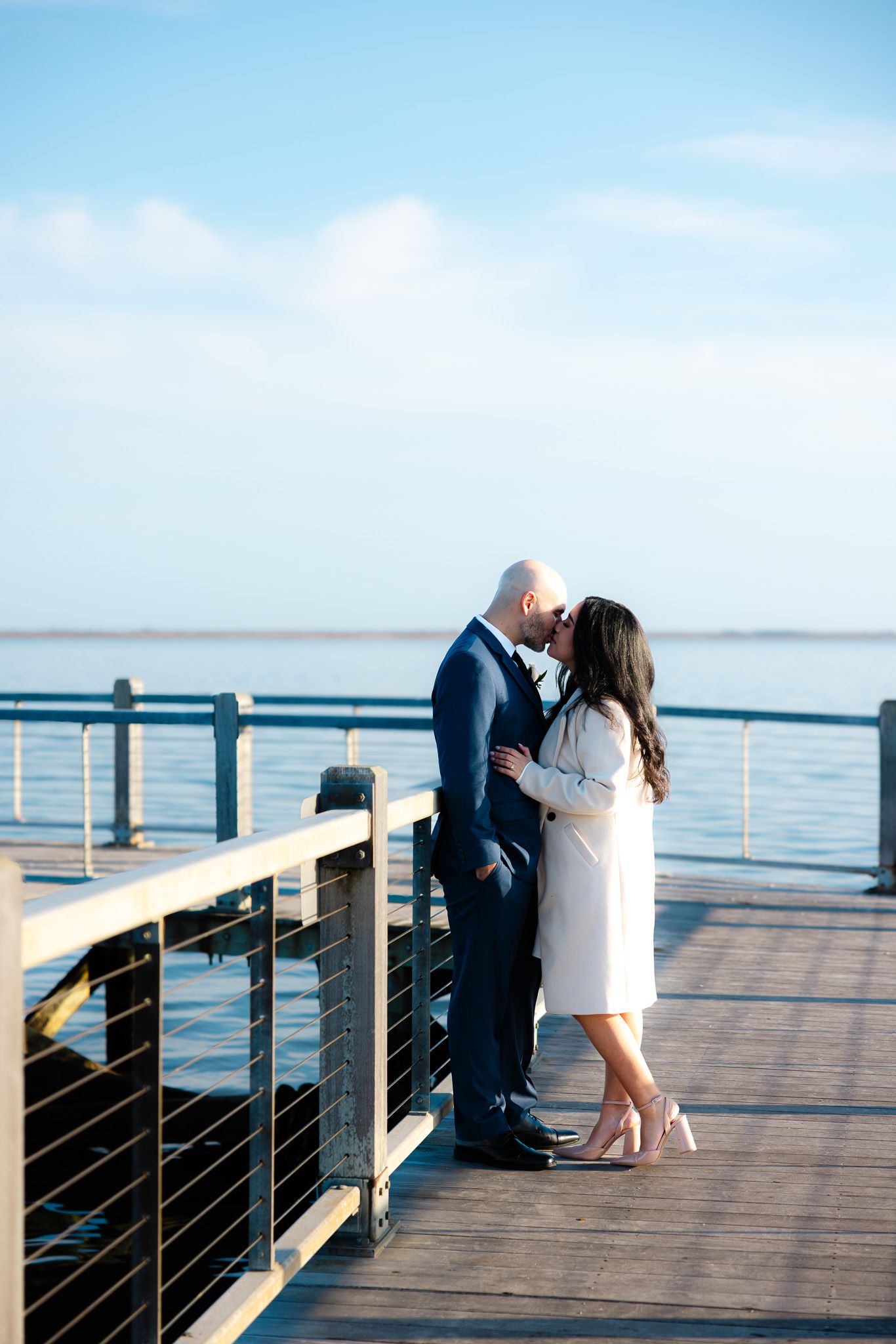 Couple sharing a romantic kiss on a waterfront pier, dressed in elegant attire with blue ocean and sky backdrop.