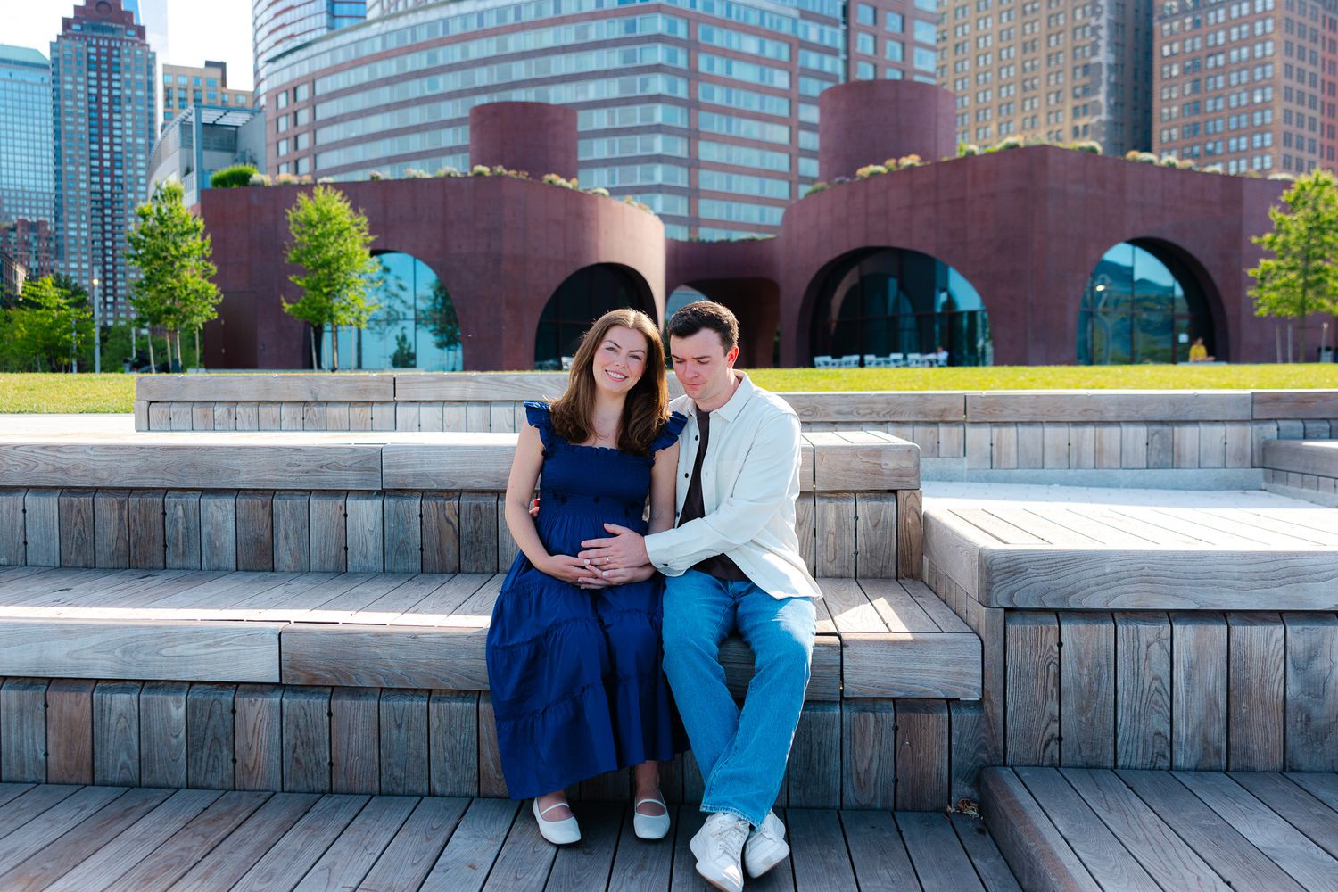 Couple sitting on wooden steps in front of modern brick building, woman in blue dress, man in white jacket, urban setting.