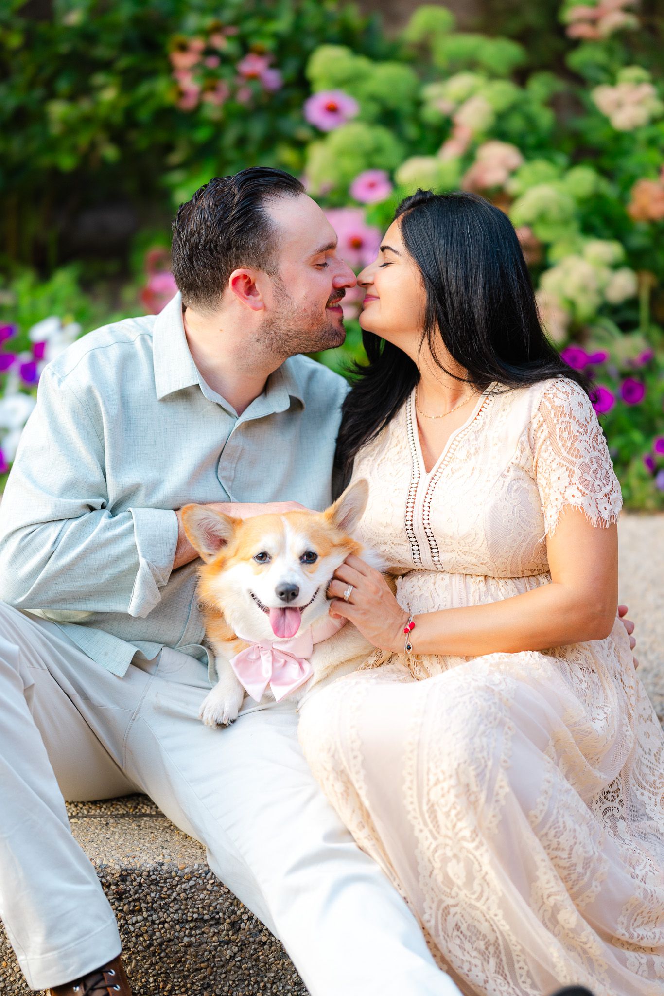 Couple sharing a romantic kiss while holding an adorable Corgi dog with a pink bow tie, seated in a flower garden.