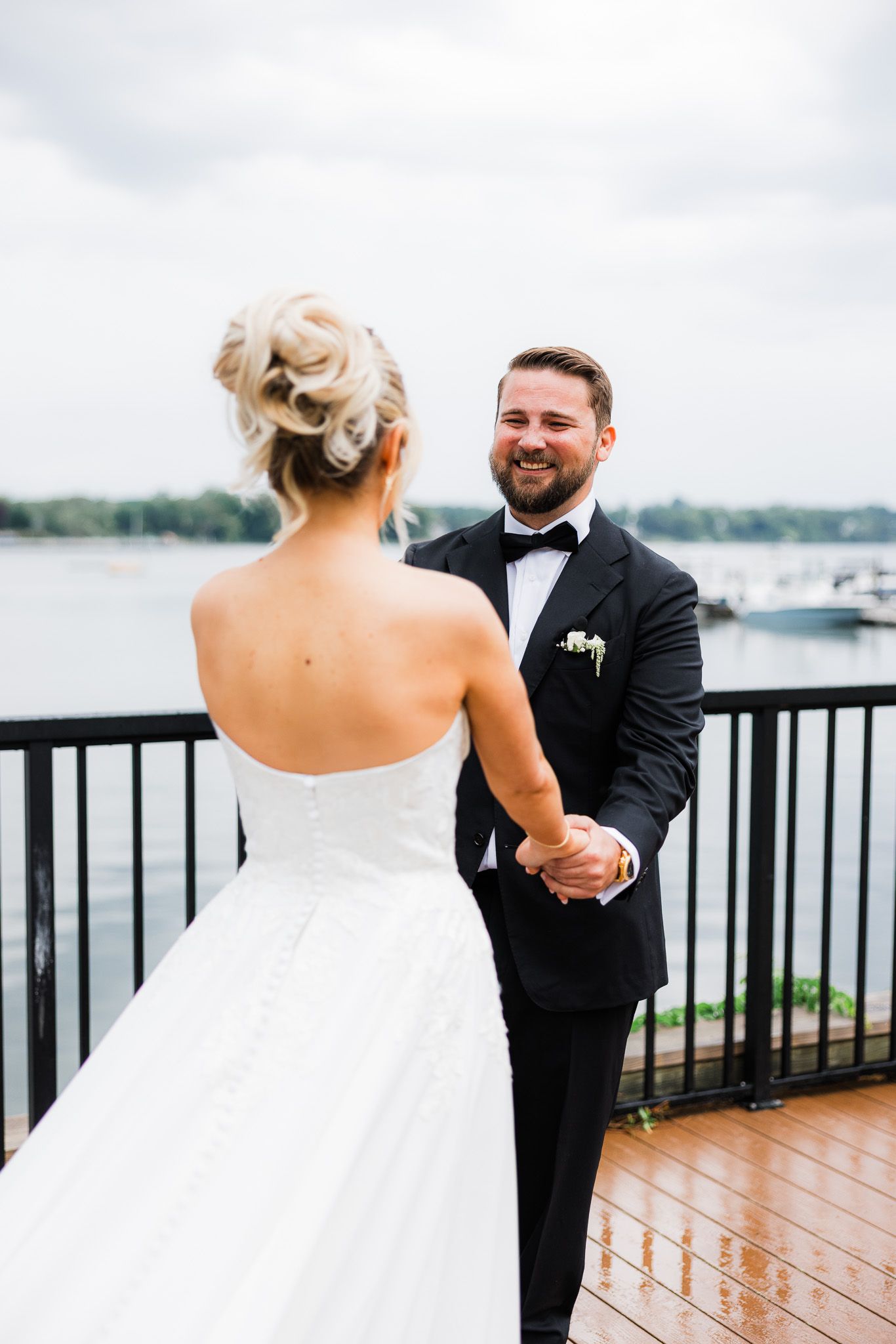 Groom in black tuxedo smiles holding bride's hands on waterfront deck at wedding.