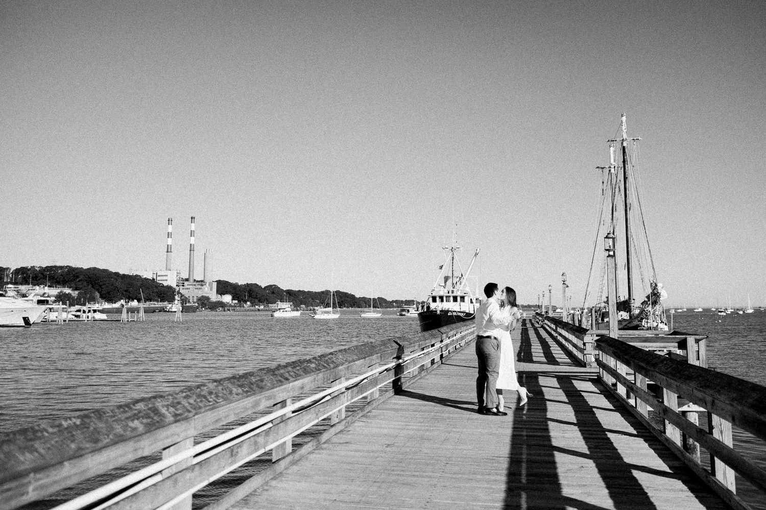 Black and white photo of a couple on a wooden pier with sailboats and calm harbor waters in the background.