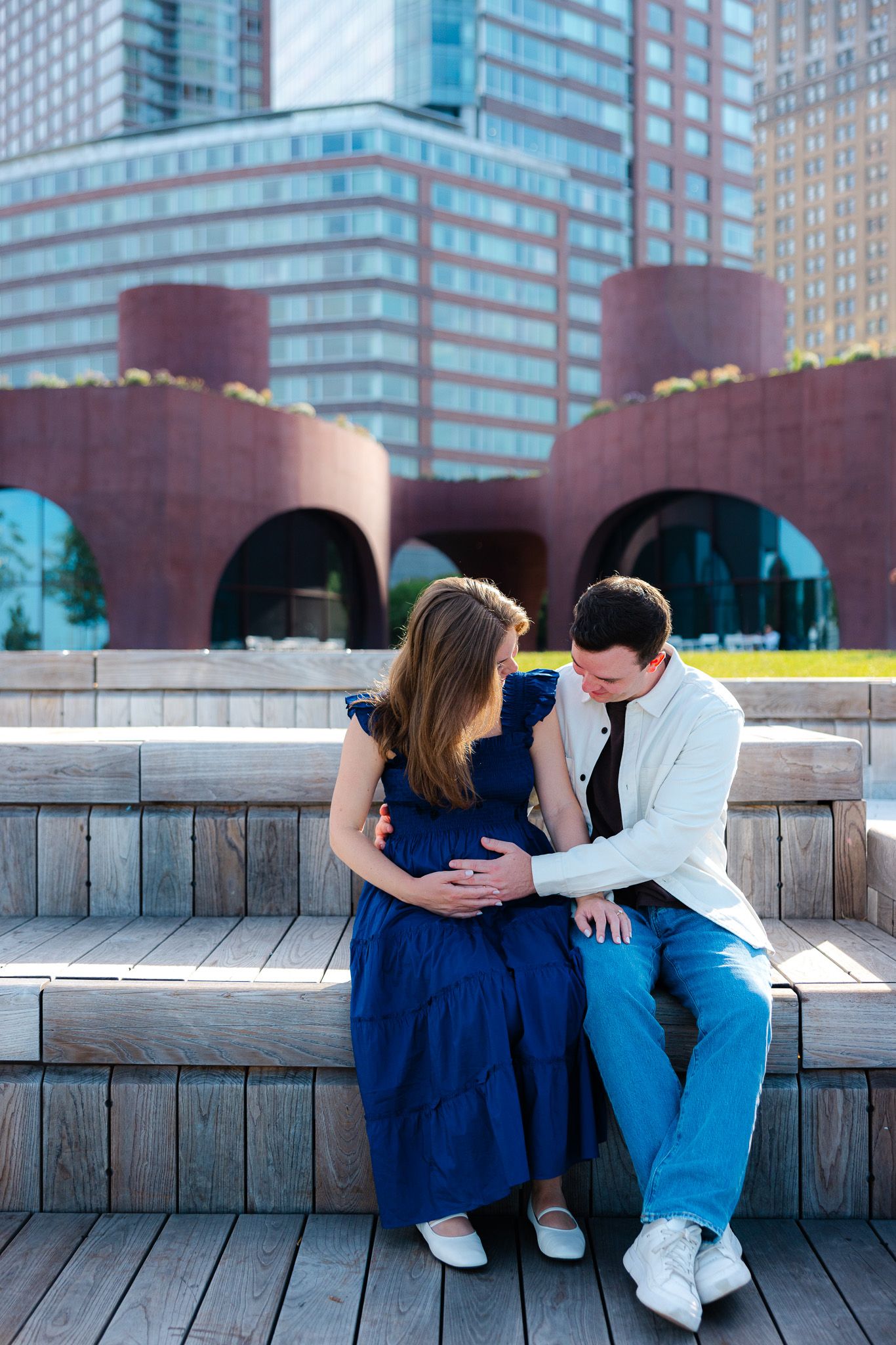 Pregnant couple sitting on wooden steps in front of modern city buildings, man tenderly touching woman's baby bump.