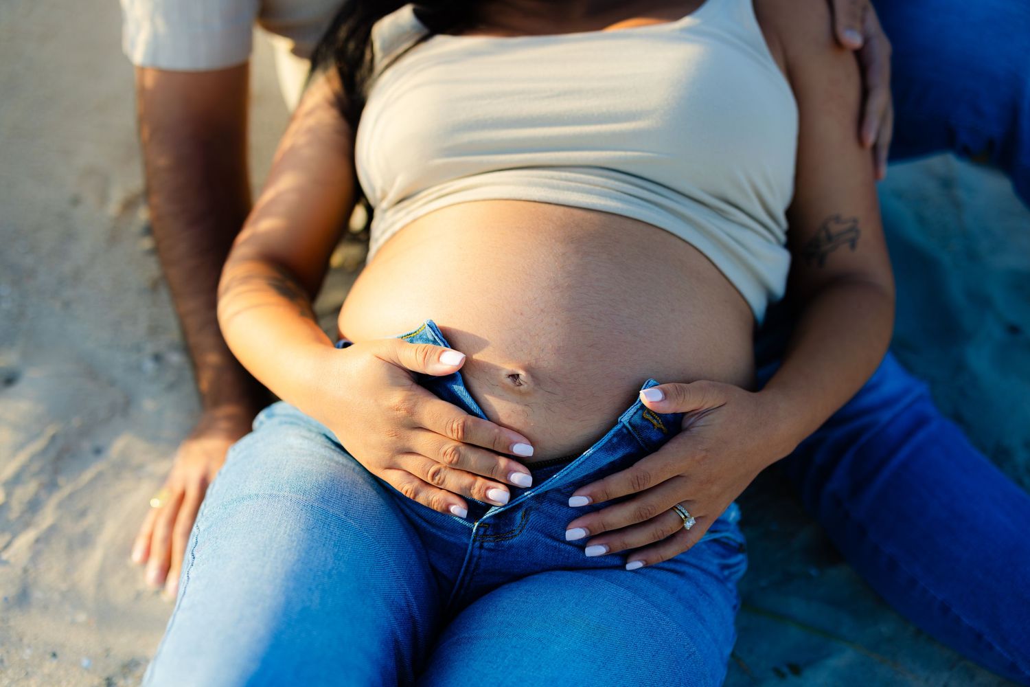 Close-up of pregnant belly with couple's hands gently cradling baby bump on sandy beach.