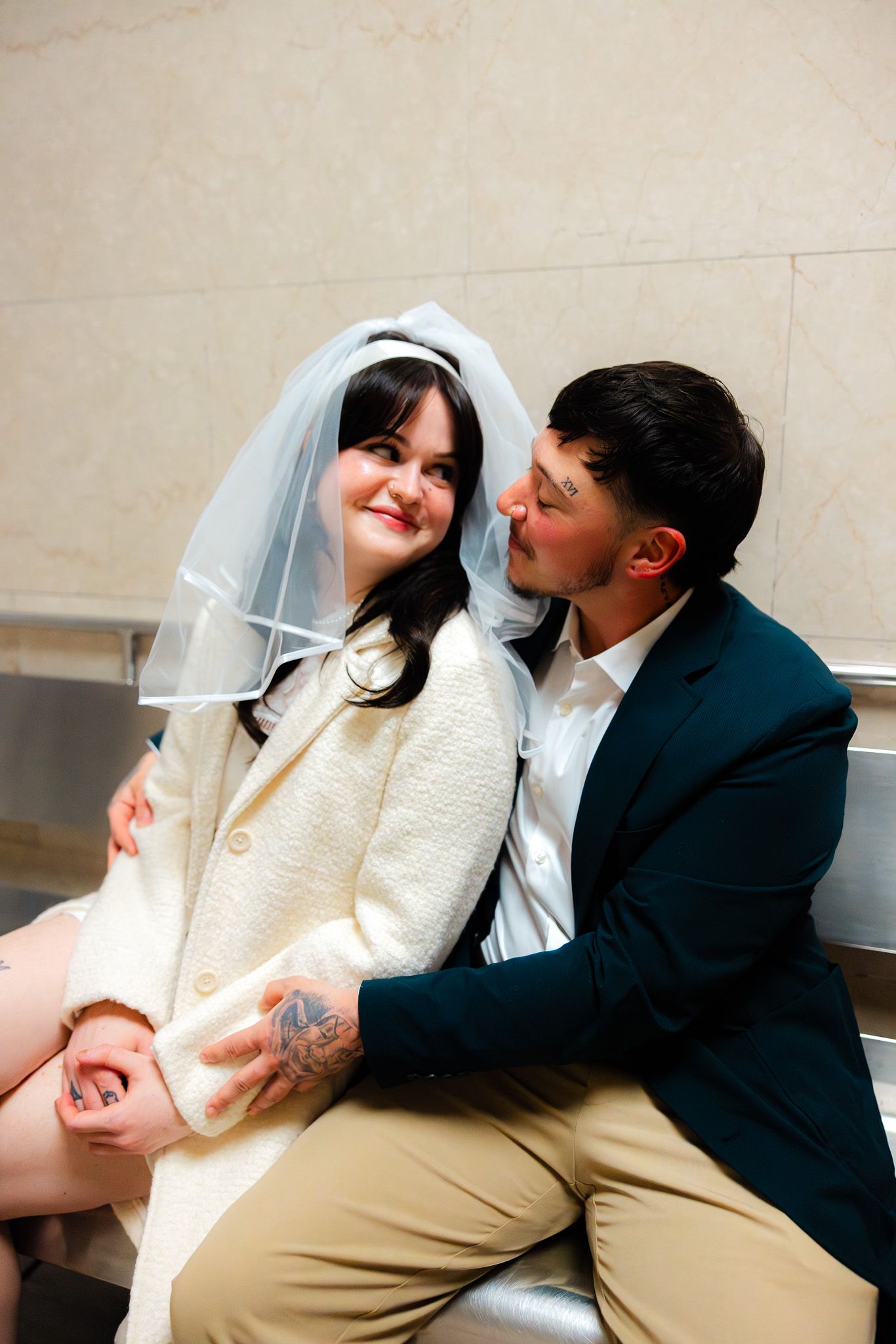 A groom in a teal suit kisses a bride wearing a white veil and coat while sitting together indoors.