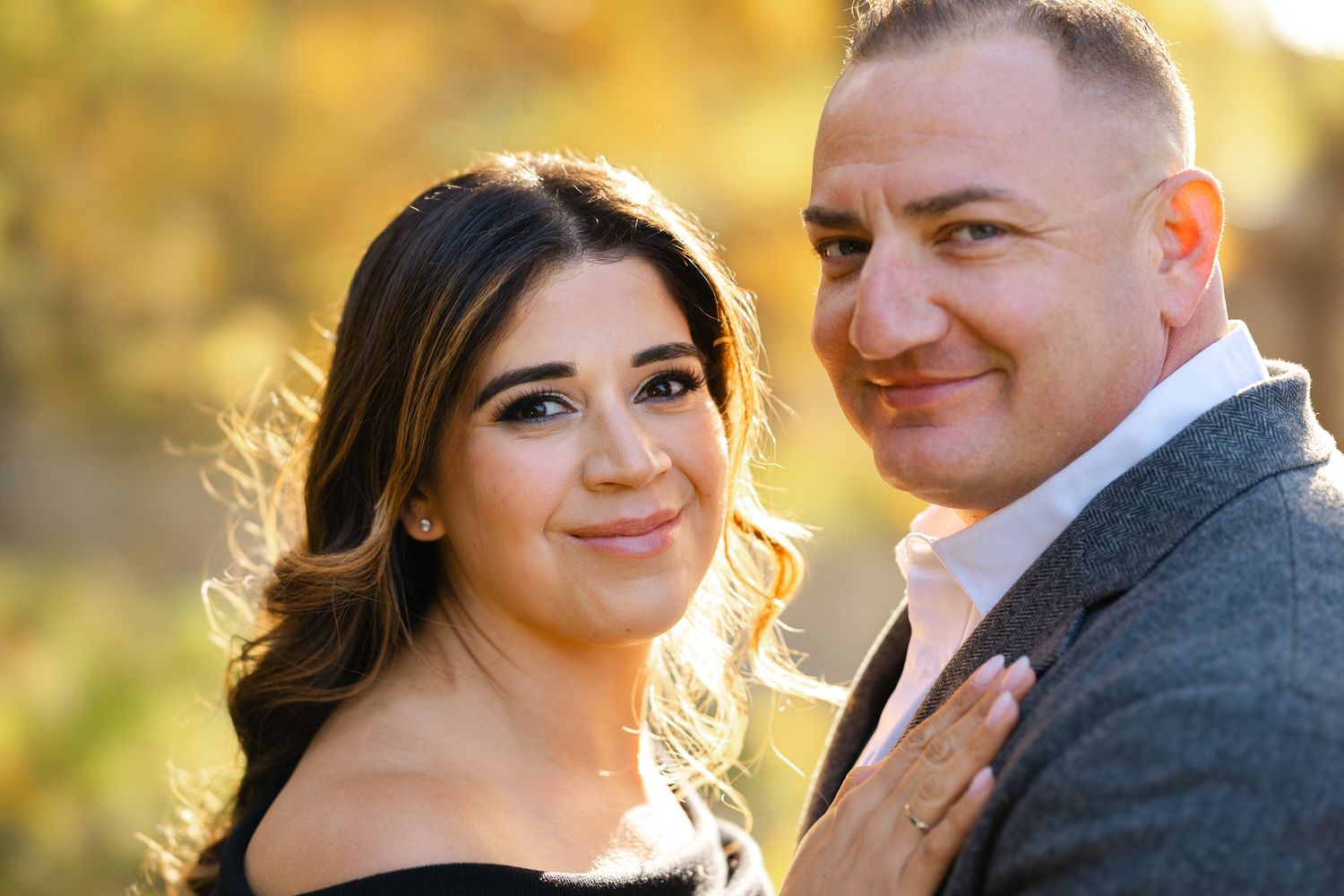 A couple poses outdoors in warm autumn light, woman in black off-shoulder top, man in gray suit jacket.