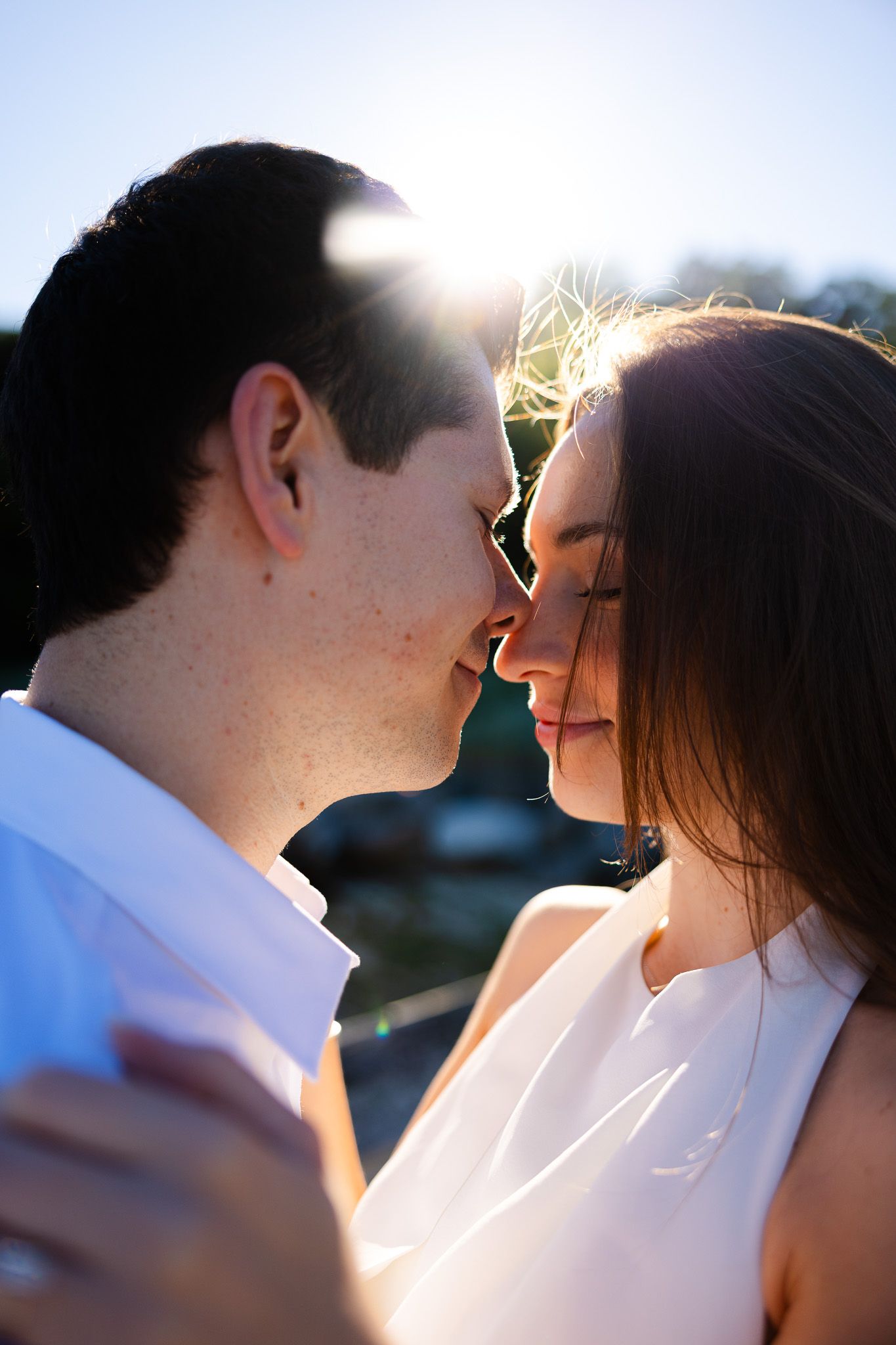 A romantic couple sharing an intimate moment, nearly kissing, backlit by golden sunlight in an outdoor setting.