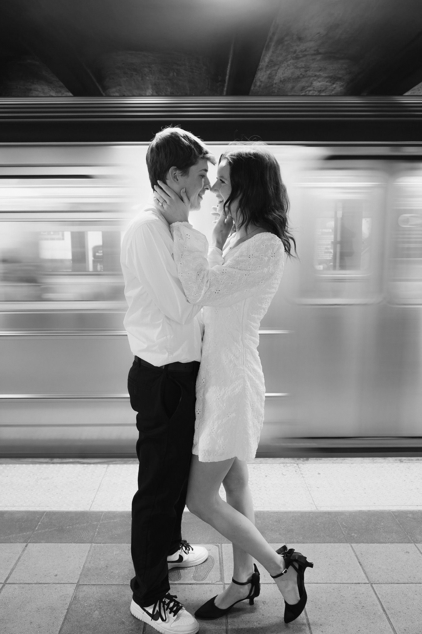 Black and white photo of couple nearly kissing on a subway platform with a speeding train behind.