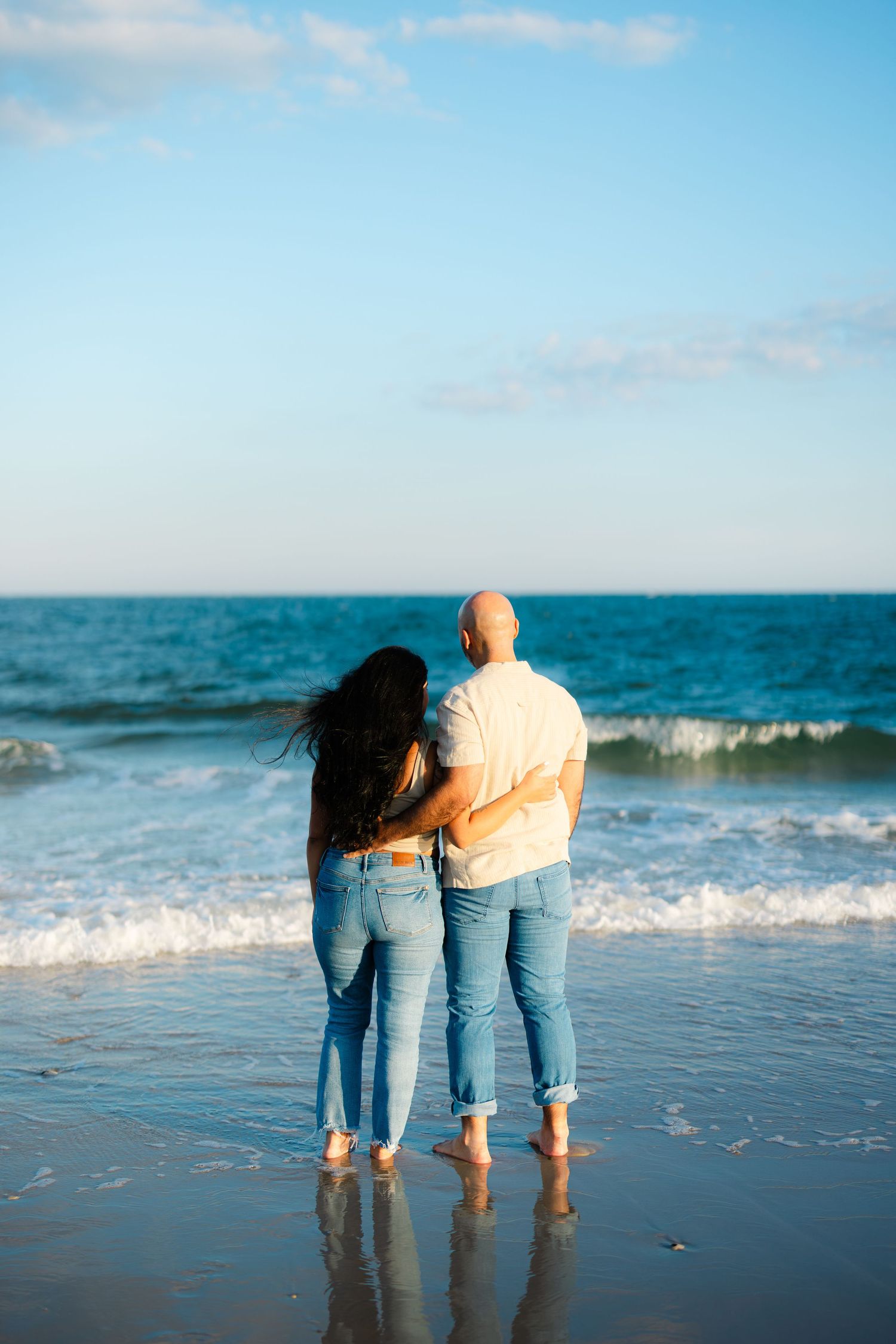 Couple embracing from behind while standing barefoot at ocean shoreline, gazing at blue waves together.