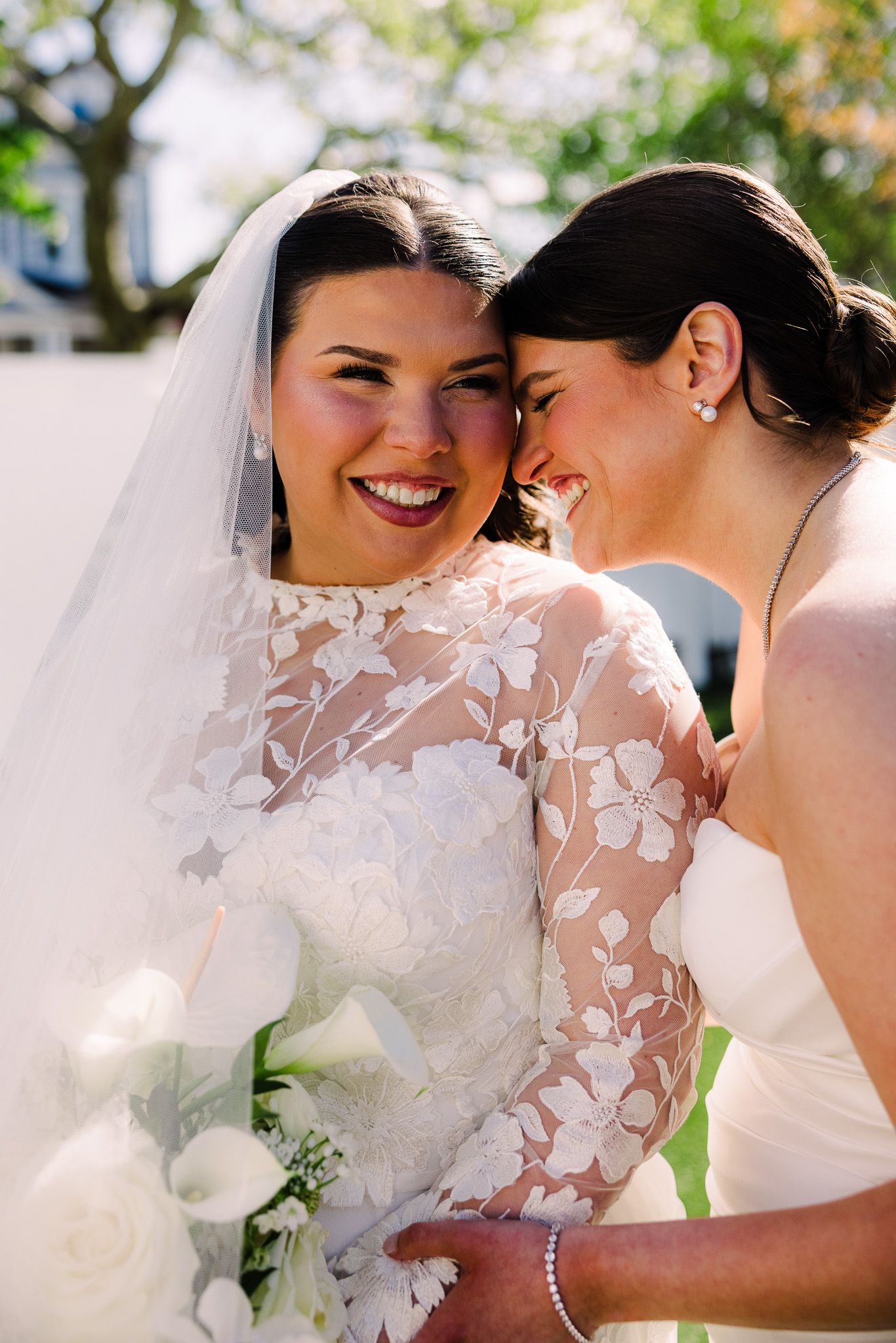 Two brides in white wedding dresses share a joyful moment, one holding a bouquet of white flowers outdoors.