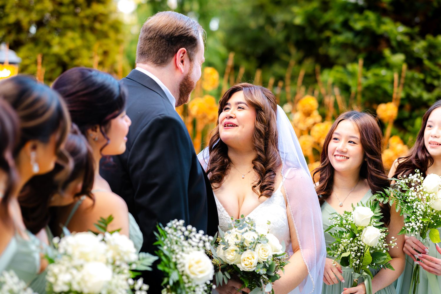 Bride smiling joyfully with groom and bridesmaids holding white floral bouquets at an outdoor wedding ceremony.