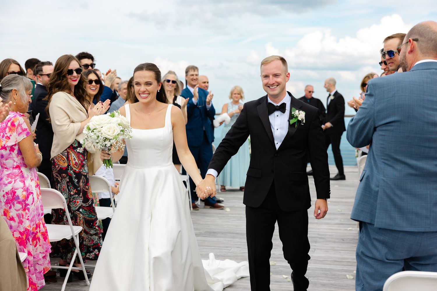 Bride and groom walk hand-in-hand down outdoor aisle on waterfront deck, guests applauding in celebration.