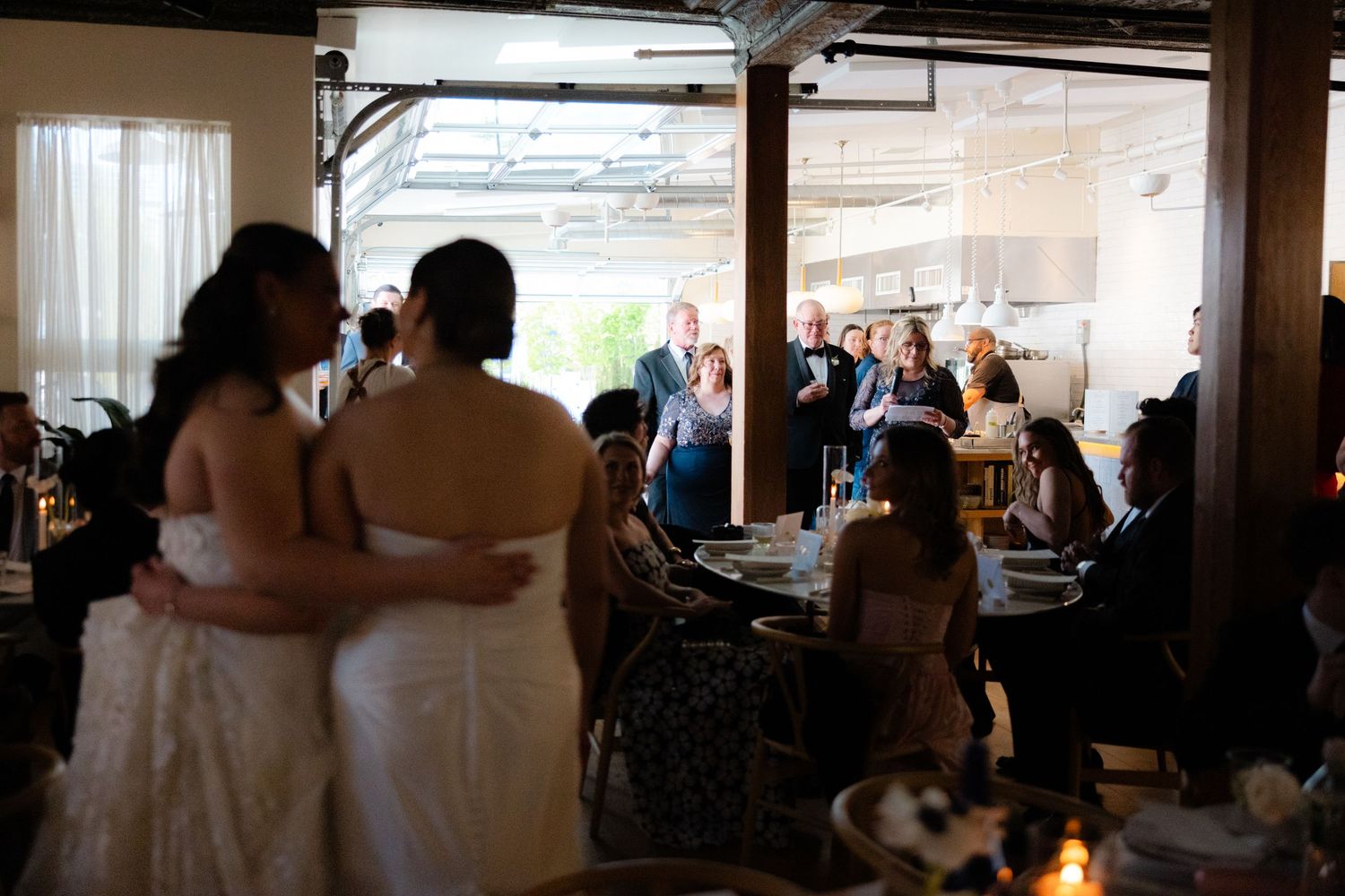 Two brides share an embrace during their wedding reception while guests celebrate around them indoors.