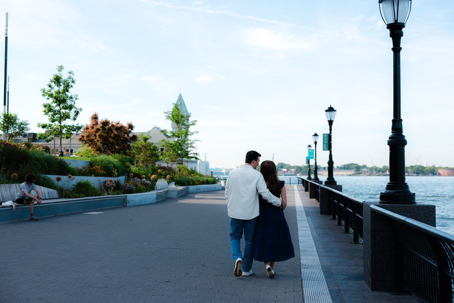 Couple walking arm in arm along a waterfront promenade with lamp posts and greenery on a clear evening.