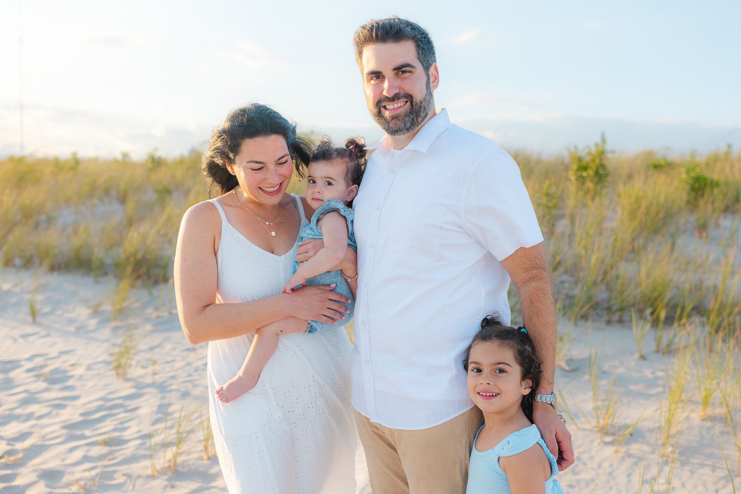 Happy family of four posing on a sandy beach, parents holding young children, wearing white and light blue outfits.