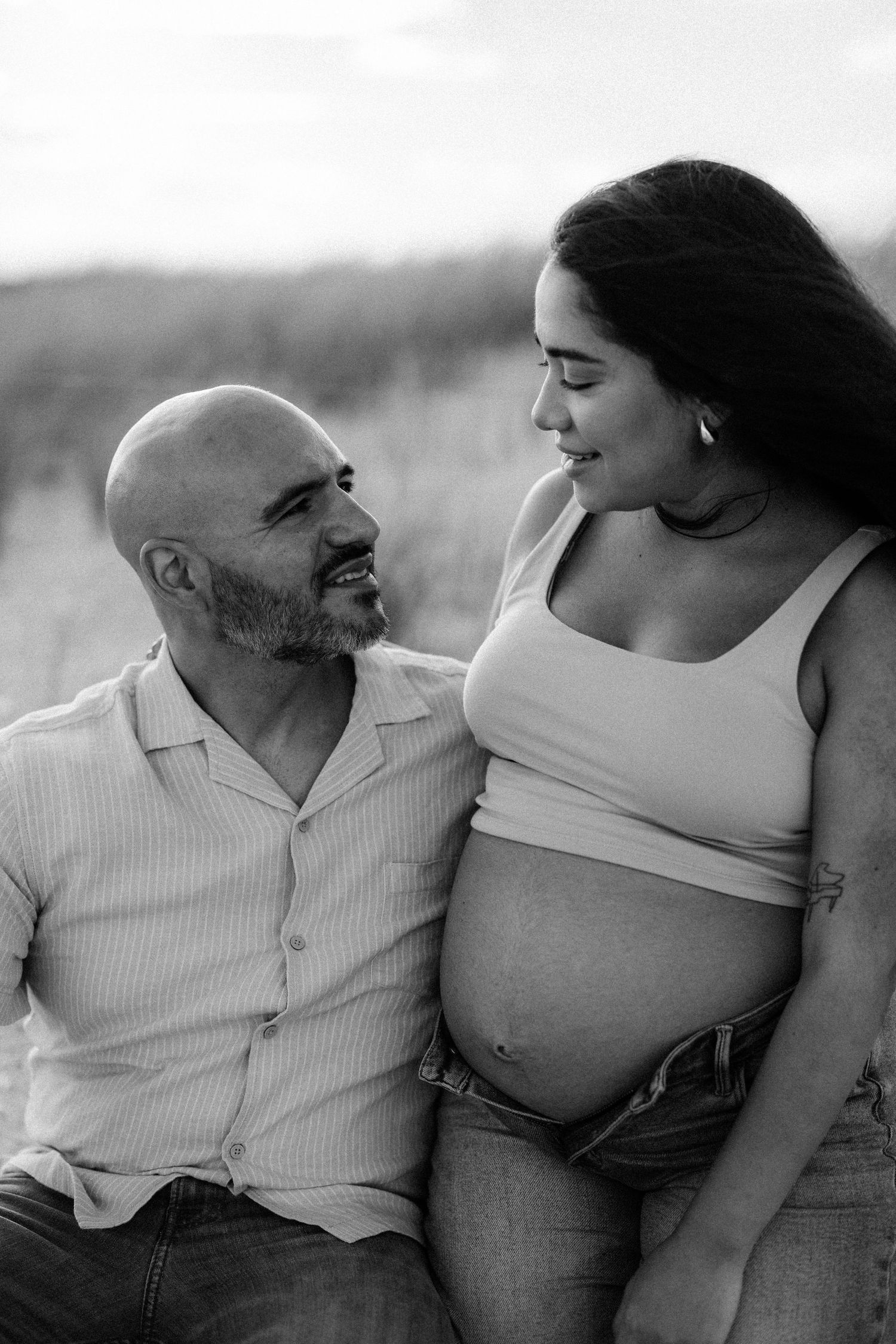 Black and white photo of a smiling pregnant couple sitting outdoors, sharing a tender moment together.