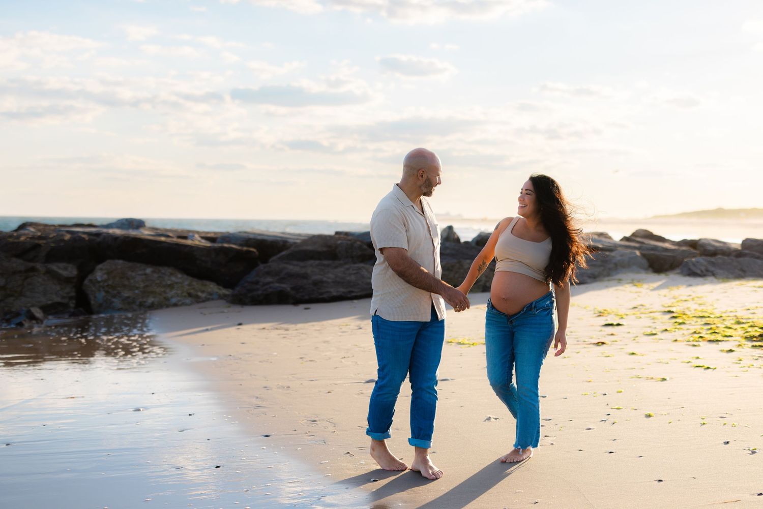 Pregnant couple holding hands on a sandy beach at sunset, with rocky shoreline and soft golden light in the background.