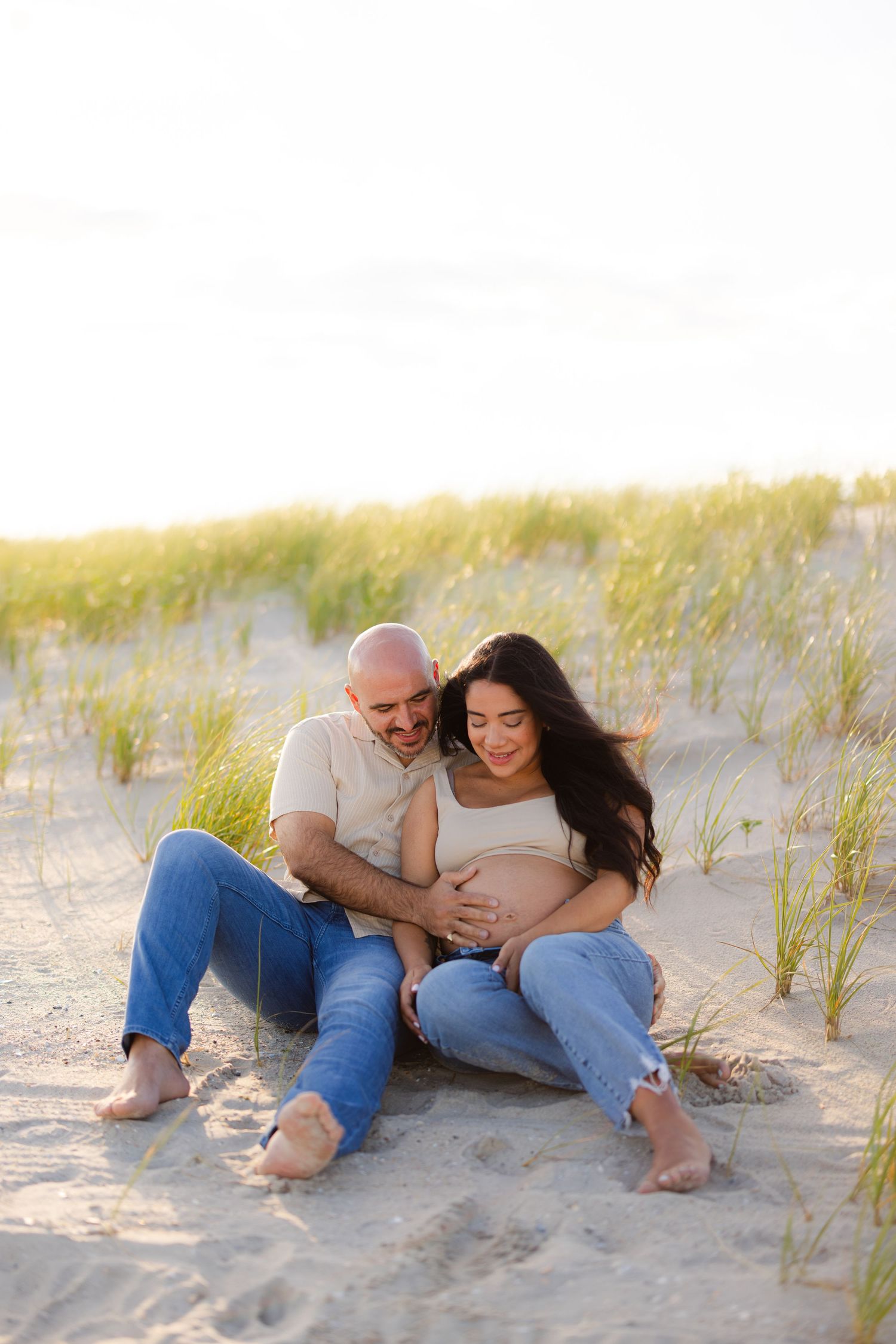 Expecting couple sitting on sandy beach dunes, smiling and holding pregnant belly in warm golden sunlight.