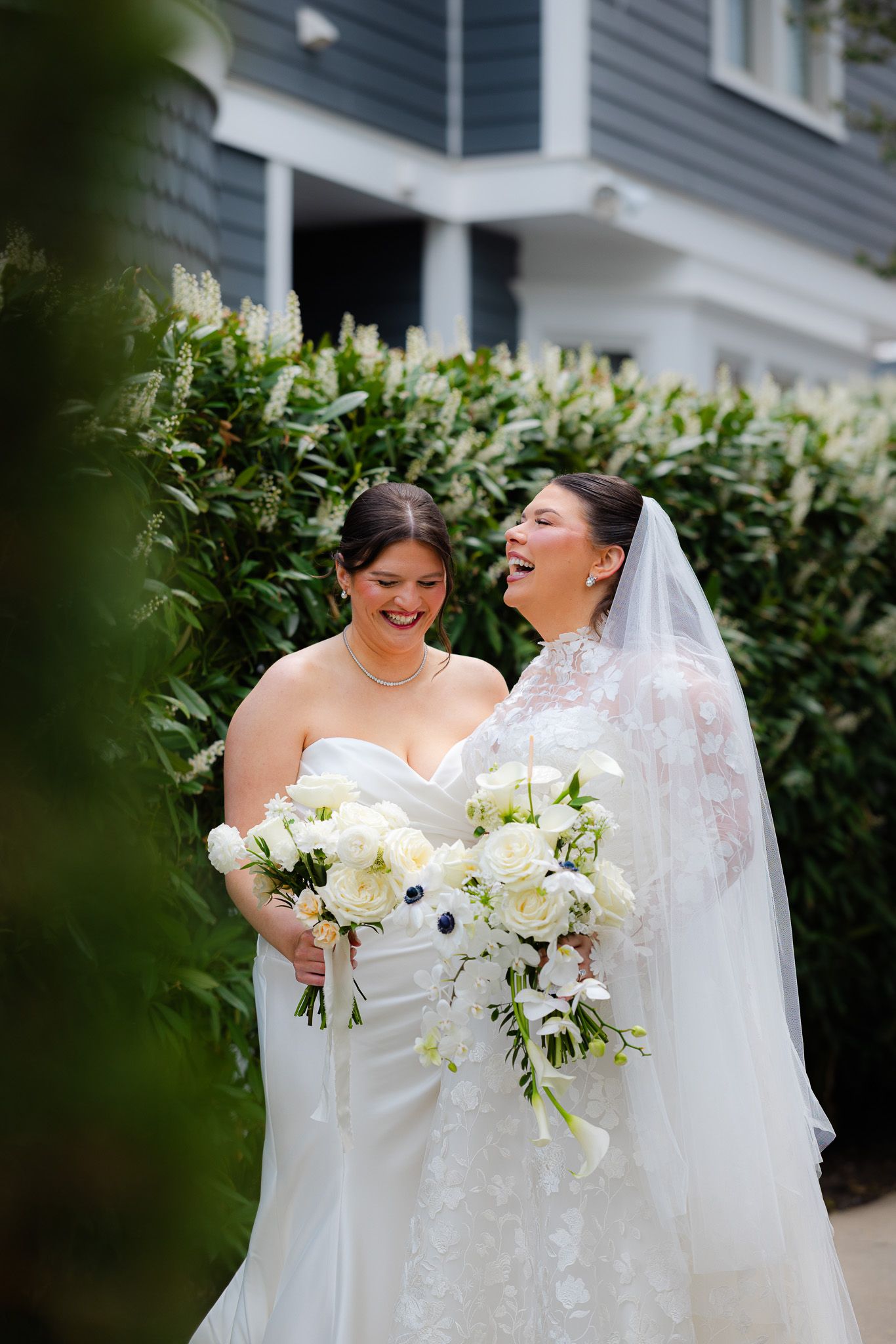 Two brides in white gowns laughing together outdoors, holding elegant white floral bouquets with greenery backdrop.