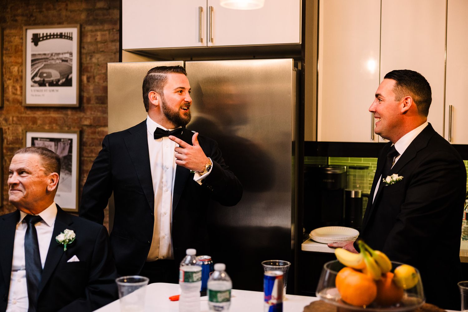 Three men in tuxedos share a laugh in a kitchen during a wedding reception celebration.
