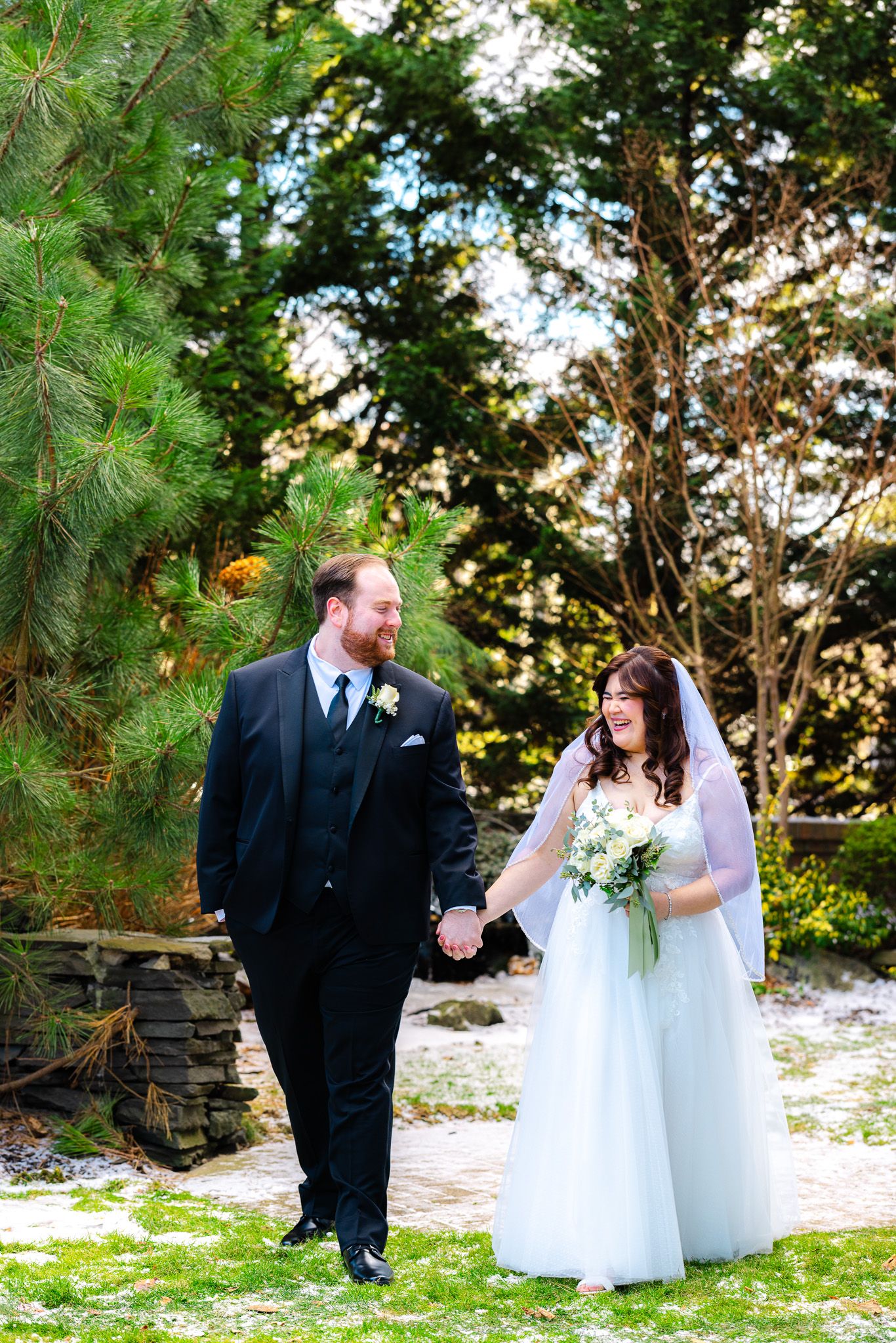 A bride and groom hold hands and smile at each other outdoors, surrounded by lush green trees on their wedding day.