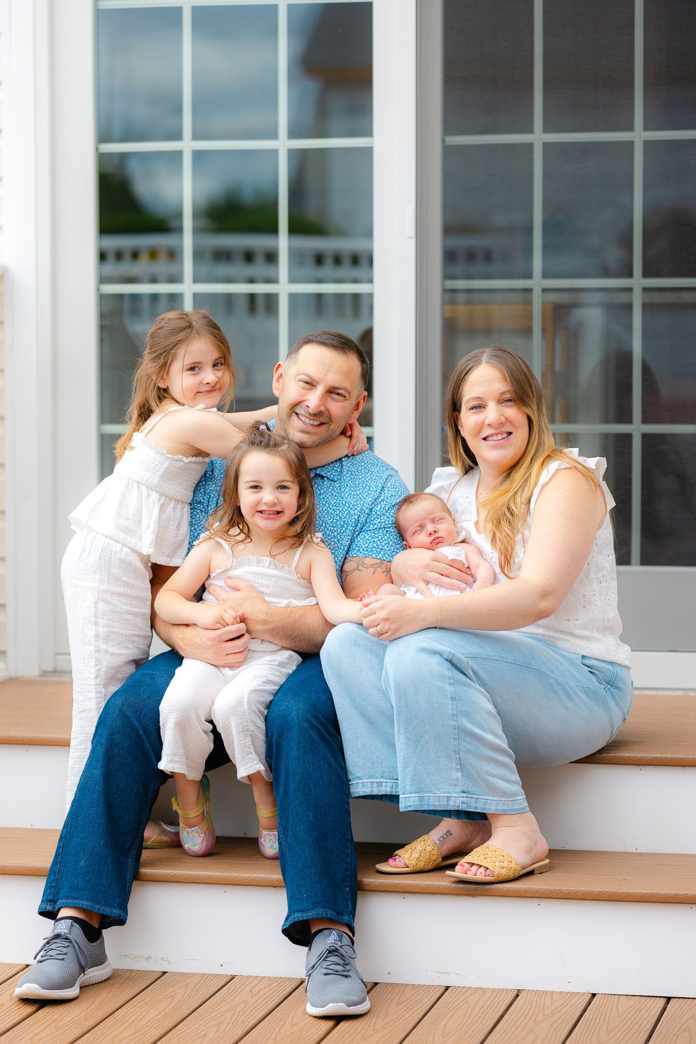 Happy family of five poses on porch steps, parents holding three young children including a newborn, smiling outdoors.