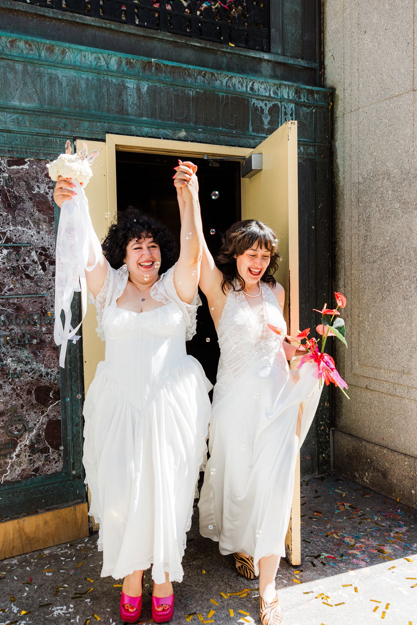 Two brides in white dresses celebrate joyfully exiting a building with raised hands and confetti.