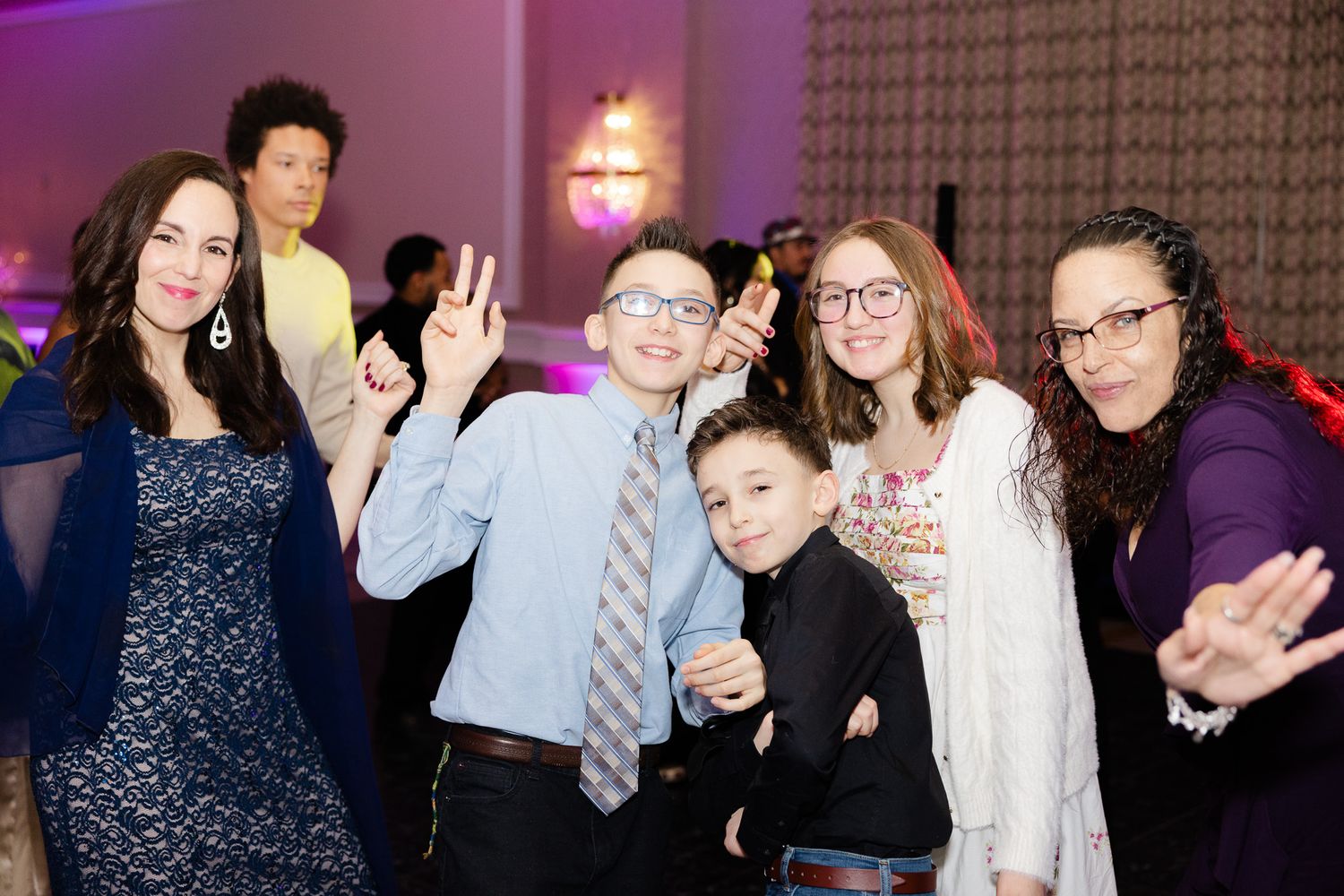 Group of joyful kids and adults posing and dancing at a formal celebration event in a decorated banquet hall.