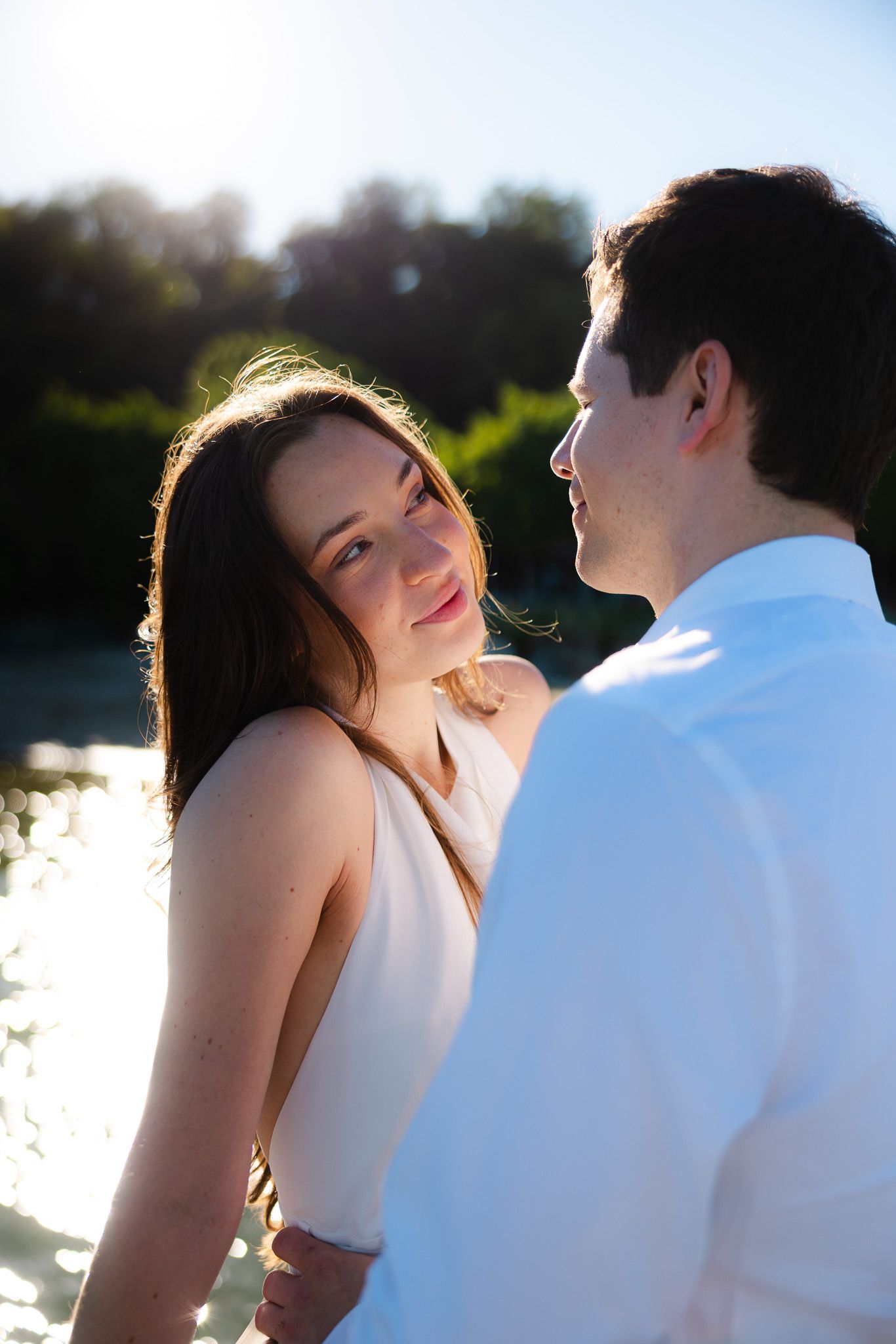 A young couple gazes at each other romantically outdoors, bathed in warm sunlight near a shimmering lake.