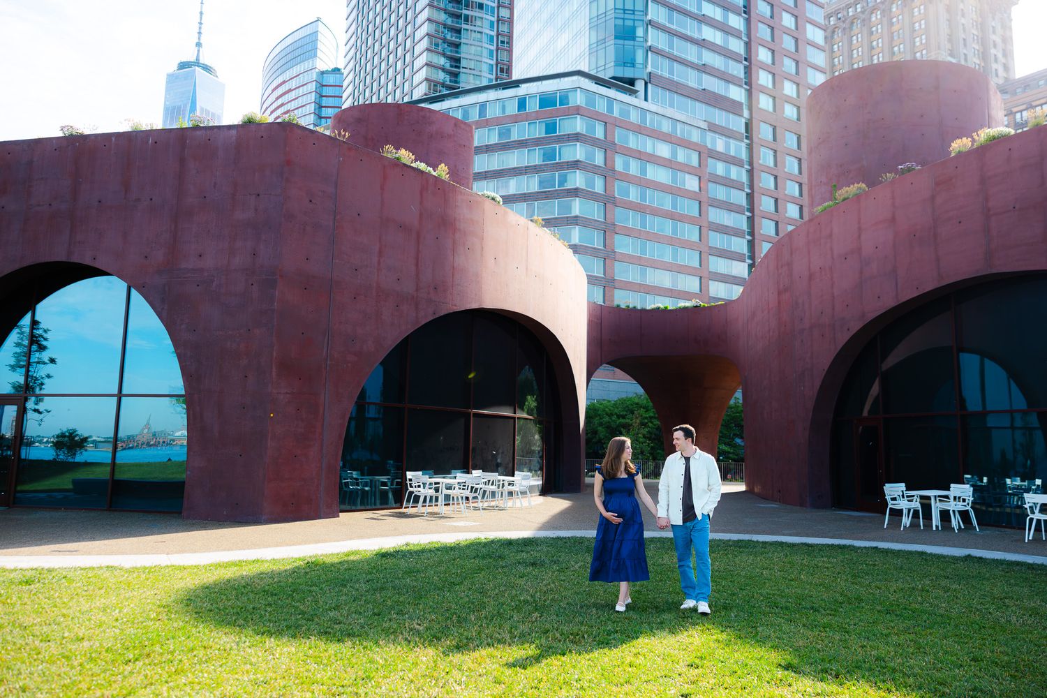 Couple holding hands on green lawn before red arched building with city skyscrapers in background.