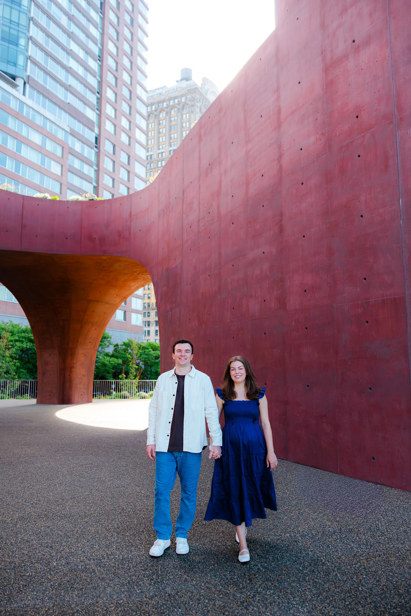 Smiling couple holding hands in front of a large red curved wall with city buildings behind them.