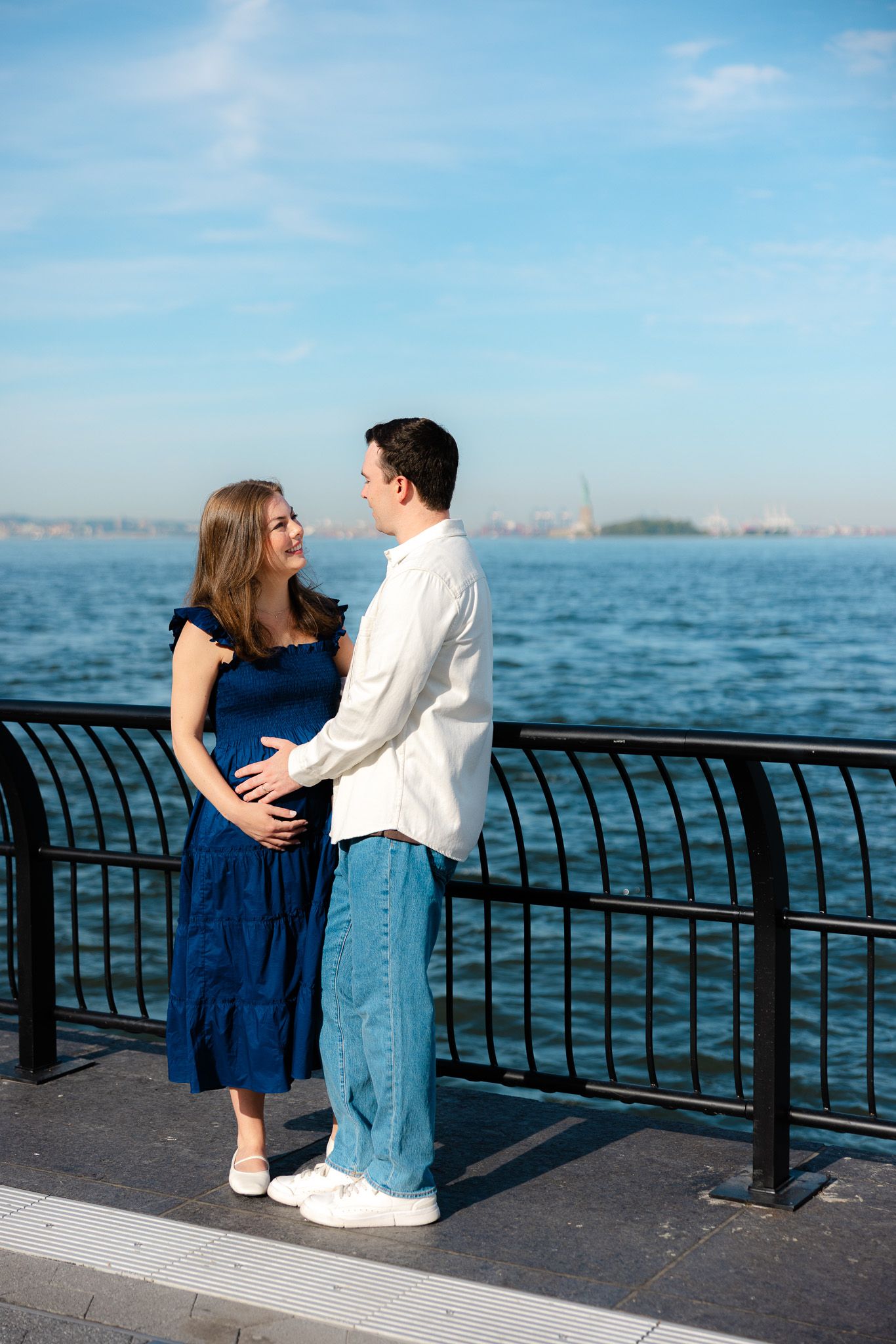 Pregnant couple embracing by waterfront railing with Statue of Liberty in background on sunny day.