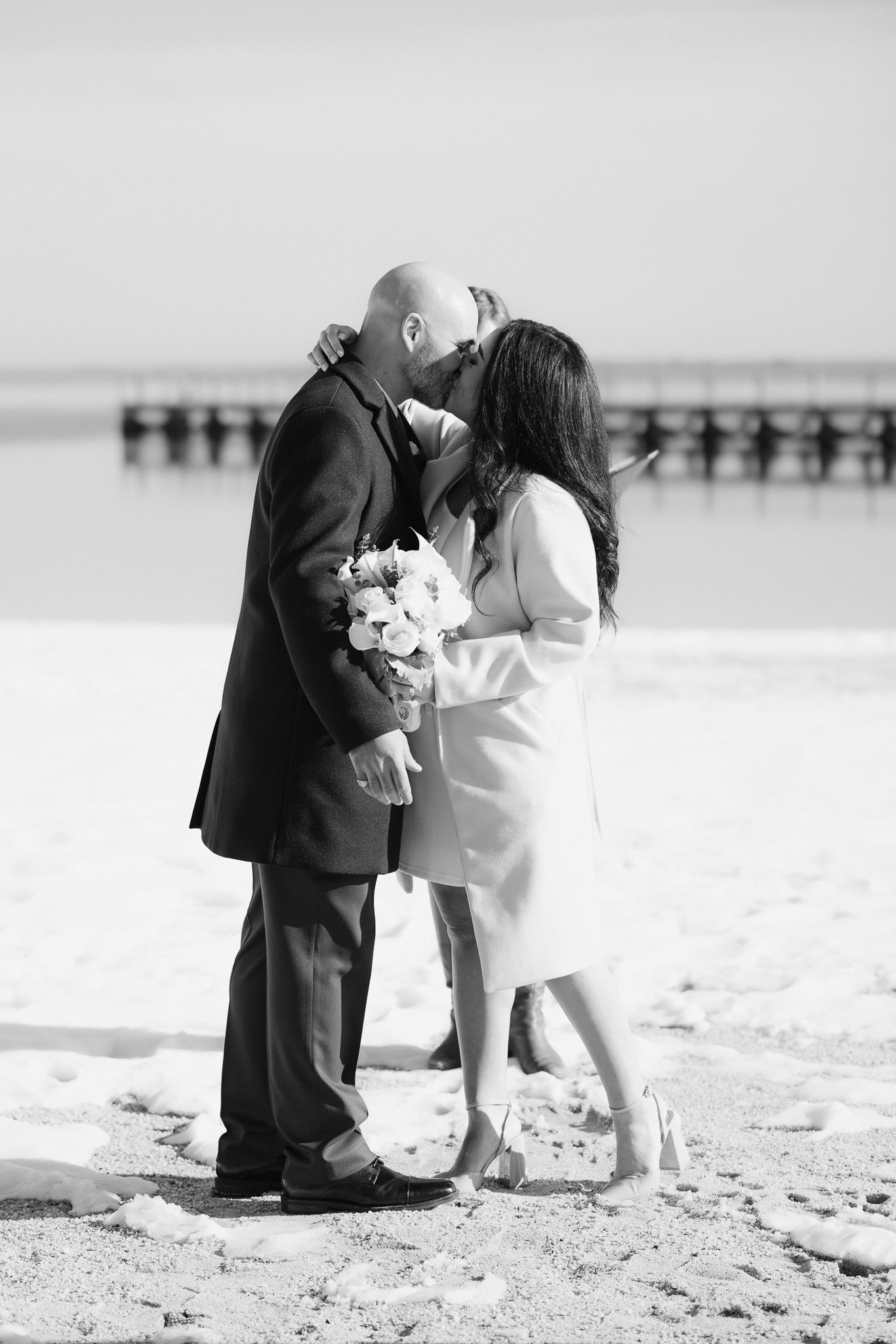 Black and white photo of a couple kissing on a snowy beach, bride holding a bouquet, with a pier in the background.