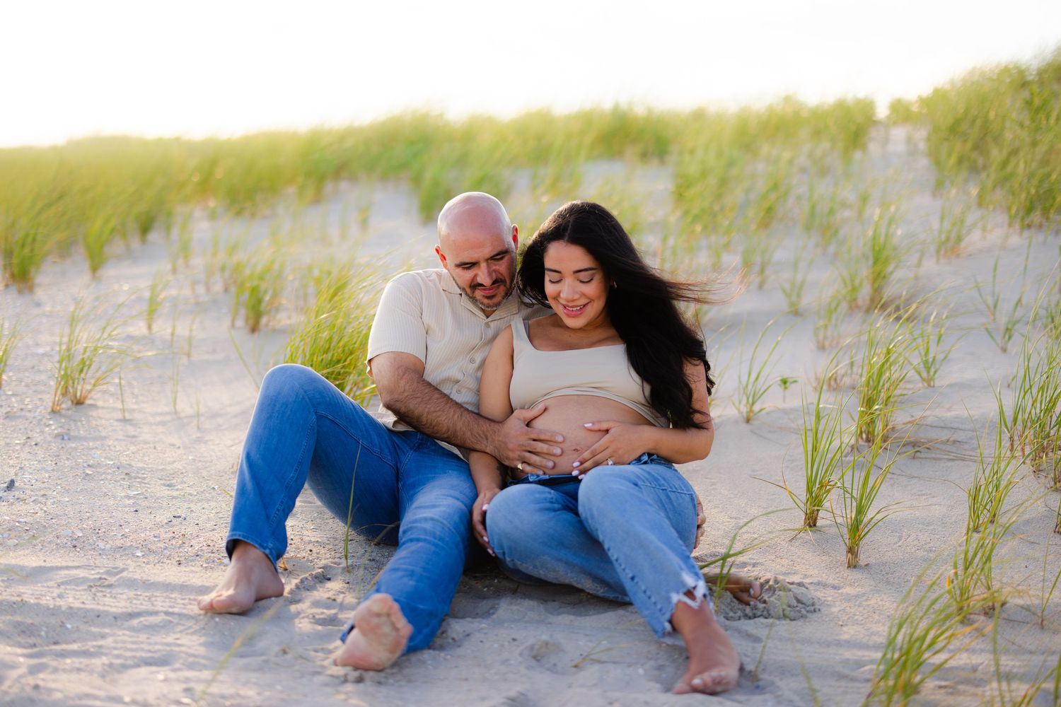 Couple sitting on sand dunes, man embracing pregnant woman while looking down at her baby bump.