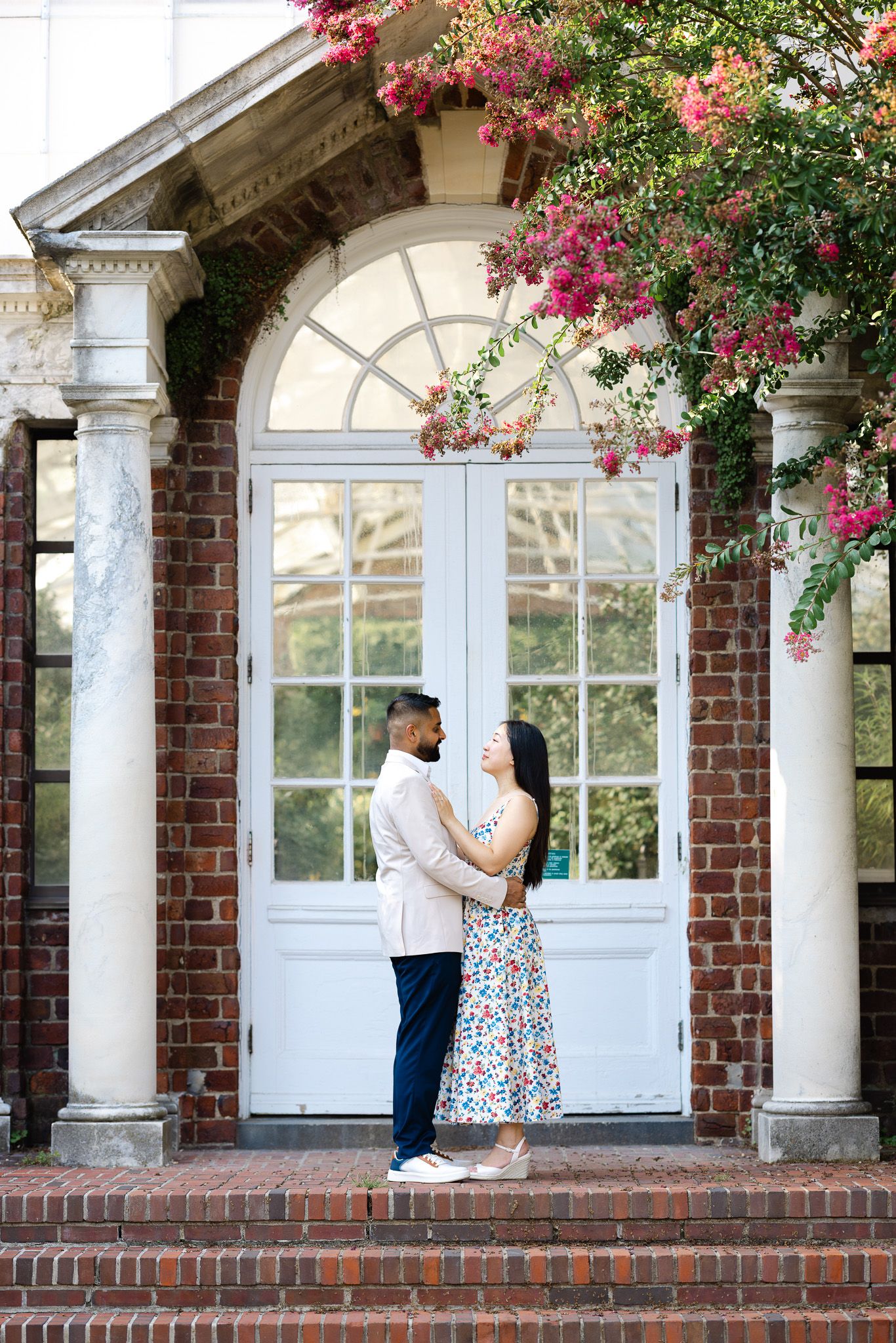 Couple embracing on brick steps before white colonial doors, framed by blooming pink crepe myrtle trees.