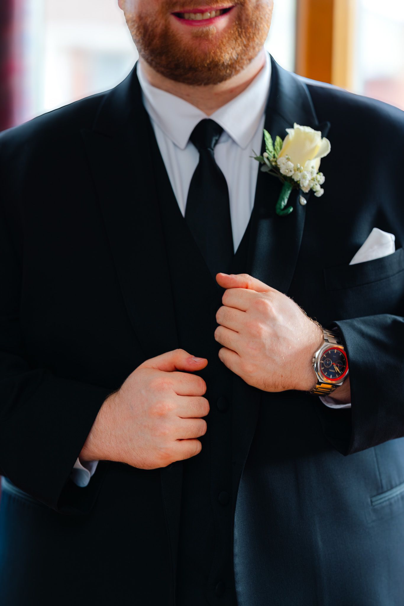 Groom in black suit adjusting jacket with white rose boutonniere and orange watch on wedding day.