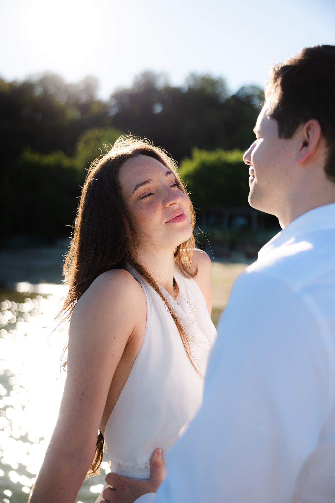 A young couple shares a romantic moment outdoors by a sunlit river, the woman smiling warmly at her partner.