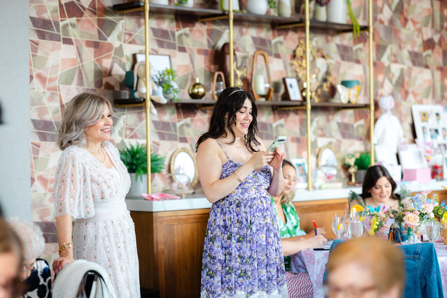 Two women in floral dresses laughing and socializing at a stylish restaurant bridal shower event.